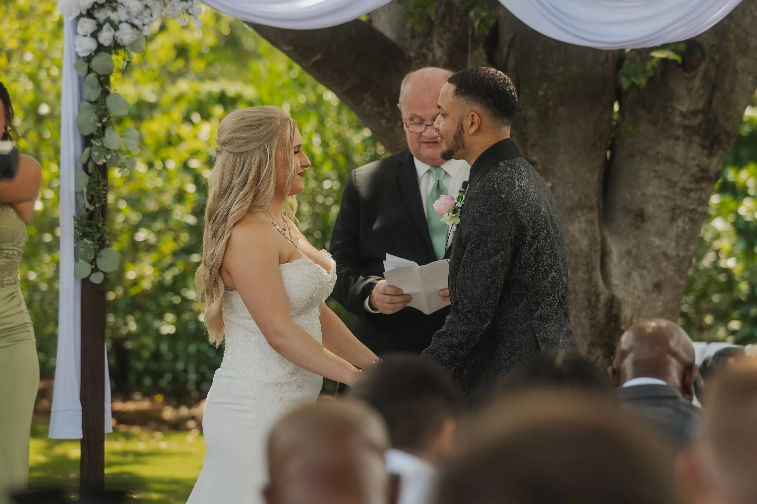 A couple getting married, standing face to face outdoors under a large tree with greenery around, with an officiant reading from papers. The bride wears a white strapless wedding gown with long wavy blonde hair, and the groom wears a dark patterned t