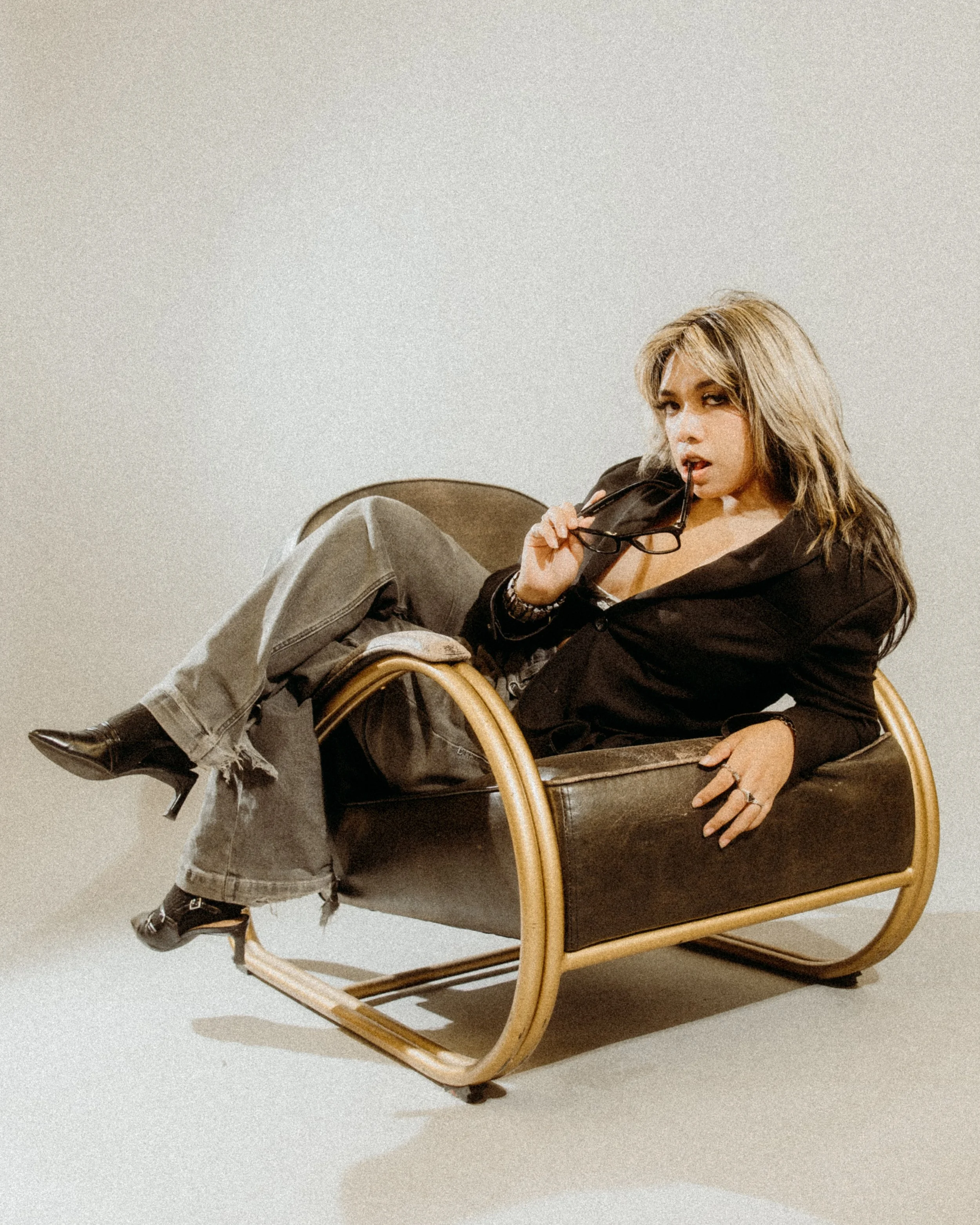 A woman with blonde hair wearing a black blazer and gray ripped jeans sits on a modern lounge chair, holding glasses and biting the arm of her glasses with a neutral expression.