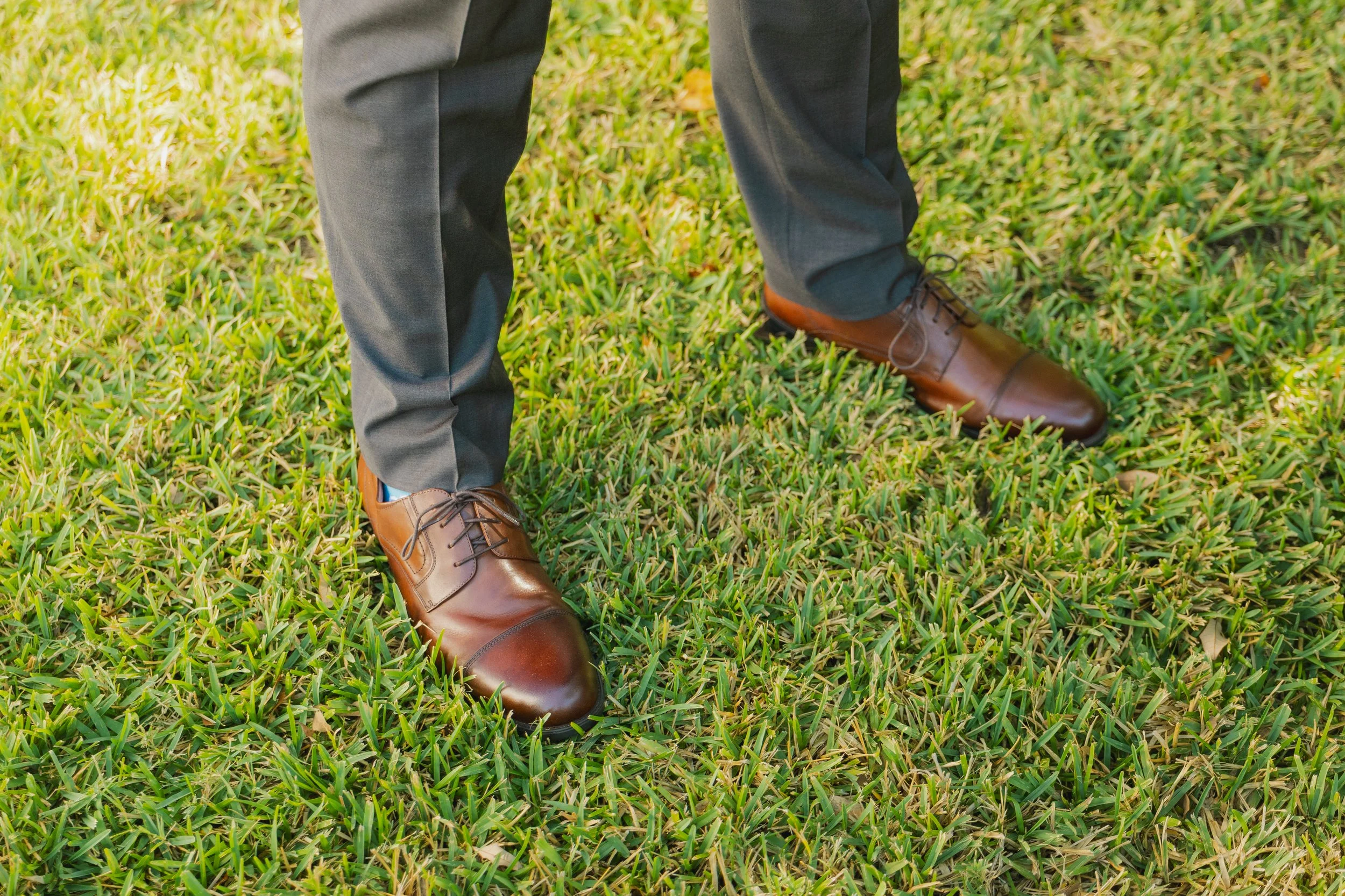 Close-up of a person's legs wearing gray slacks and brown leather dress shoes standing on green grass.
