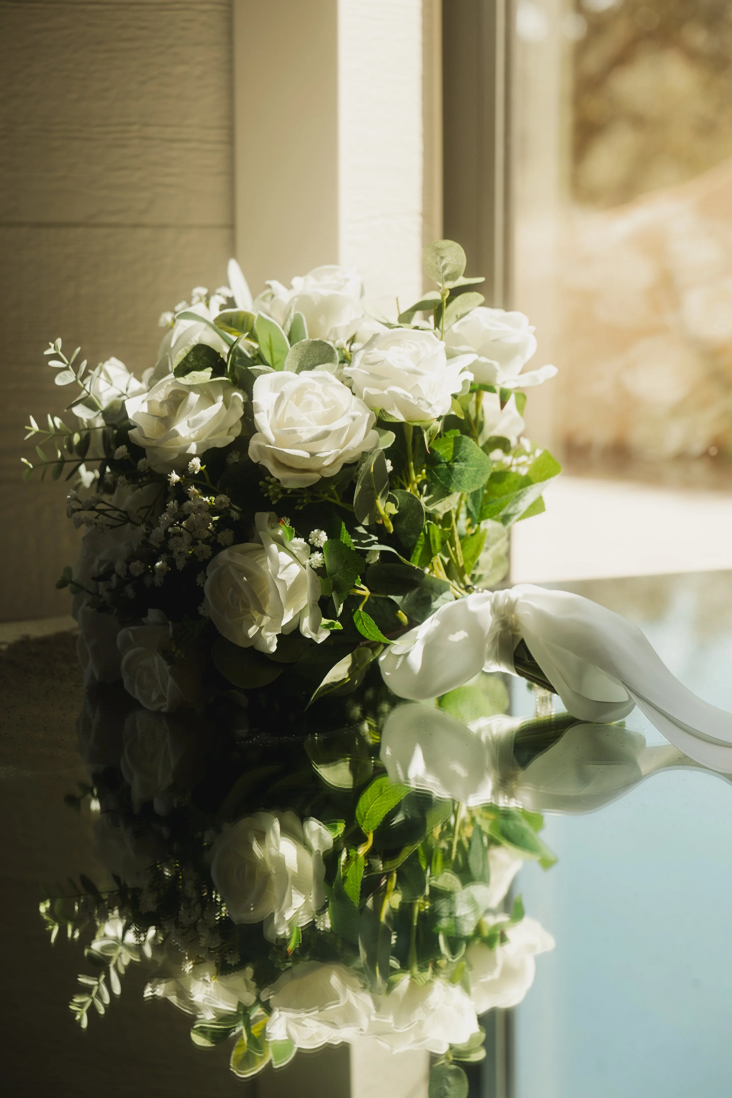 A bouquet of white roses and greenery with a white ribbon, resting on a reflective surface near a window letting in natural light.