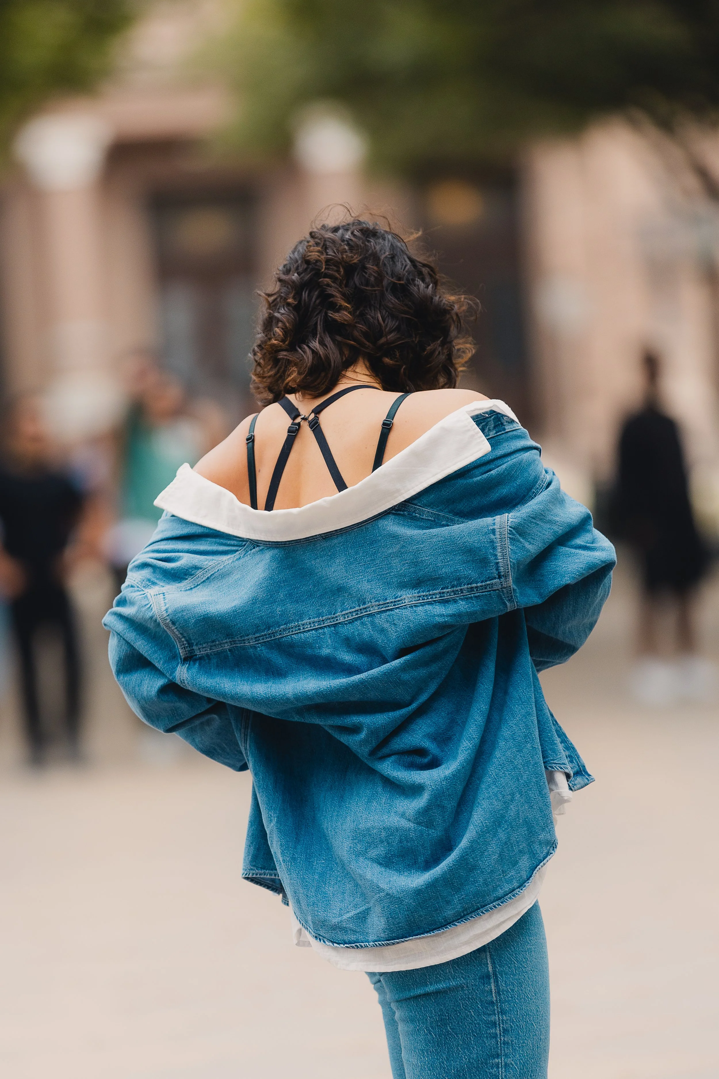 Back view of a woman adjusting her denim jacket with curly hair, standing outdoors with a blurred background of people and buildings.