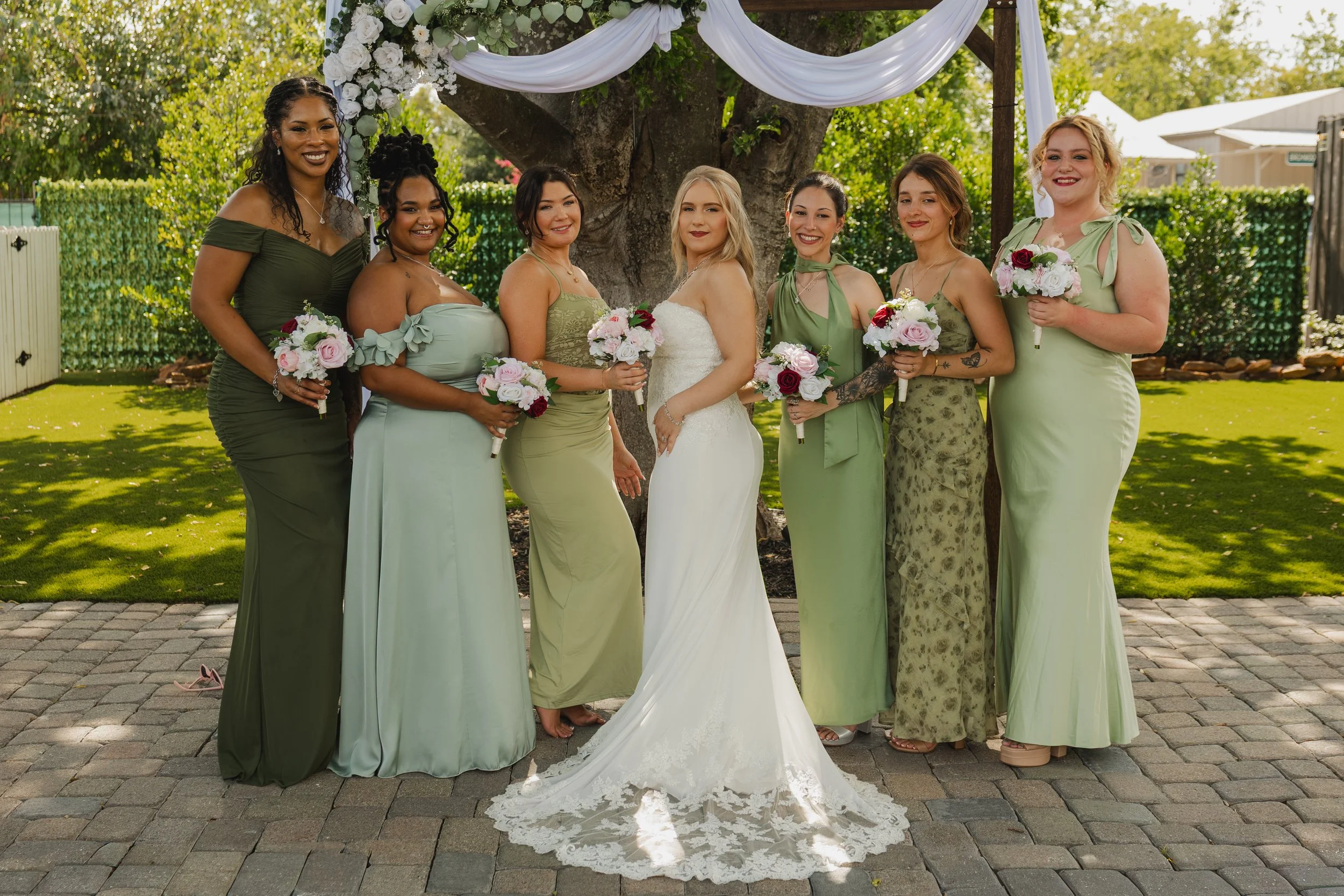 A group of nine women, including a bride in a white wedding dress, standing outdoors under a large tree decorated with white fabric and flowers. They are all holding small bouquets of pink and white flowers, and are dressed in various shades of green