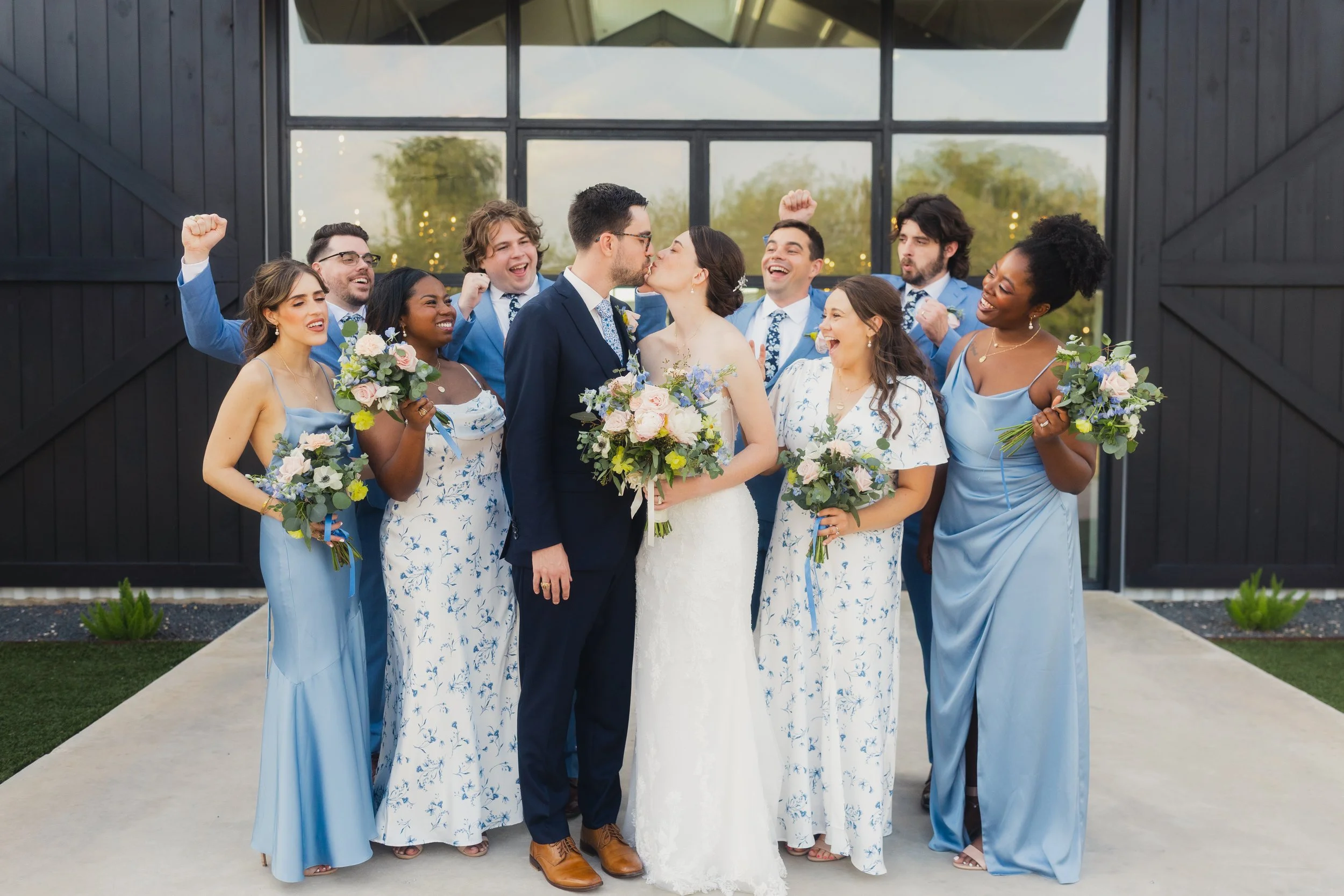 A wedding celebration with a bride and groom sharing a kiss, surrounded by bridesmaids and groomsmen, outdoors with a modern dark barn-style building in the background.