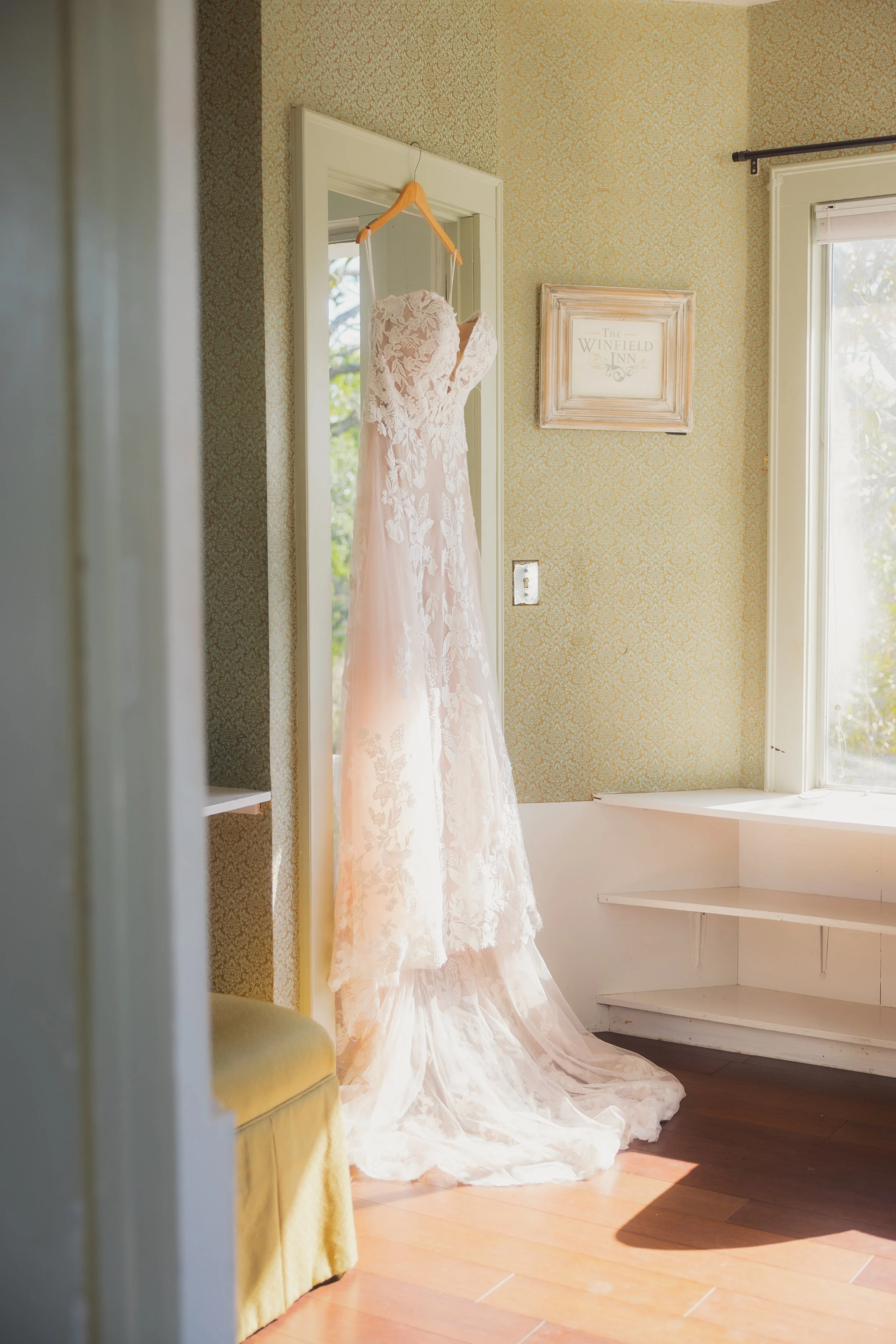A wedding dress hanging on a wooden hanger on a door in a sunlit room with vintage wallpaper and a picture frame on the wall.