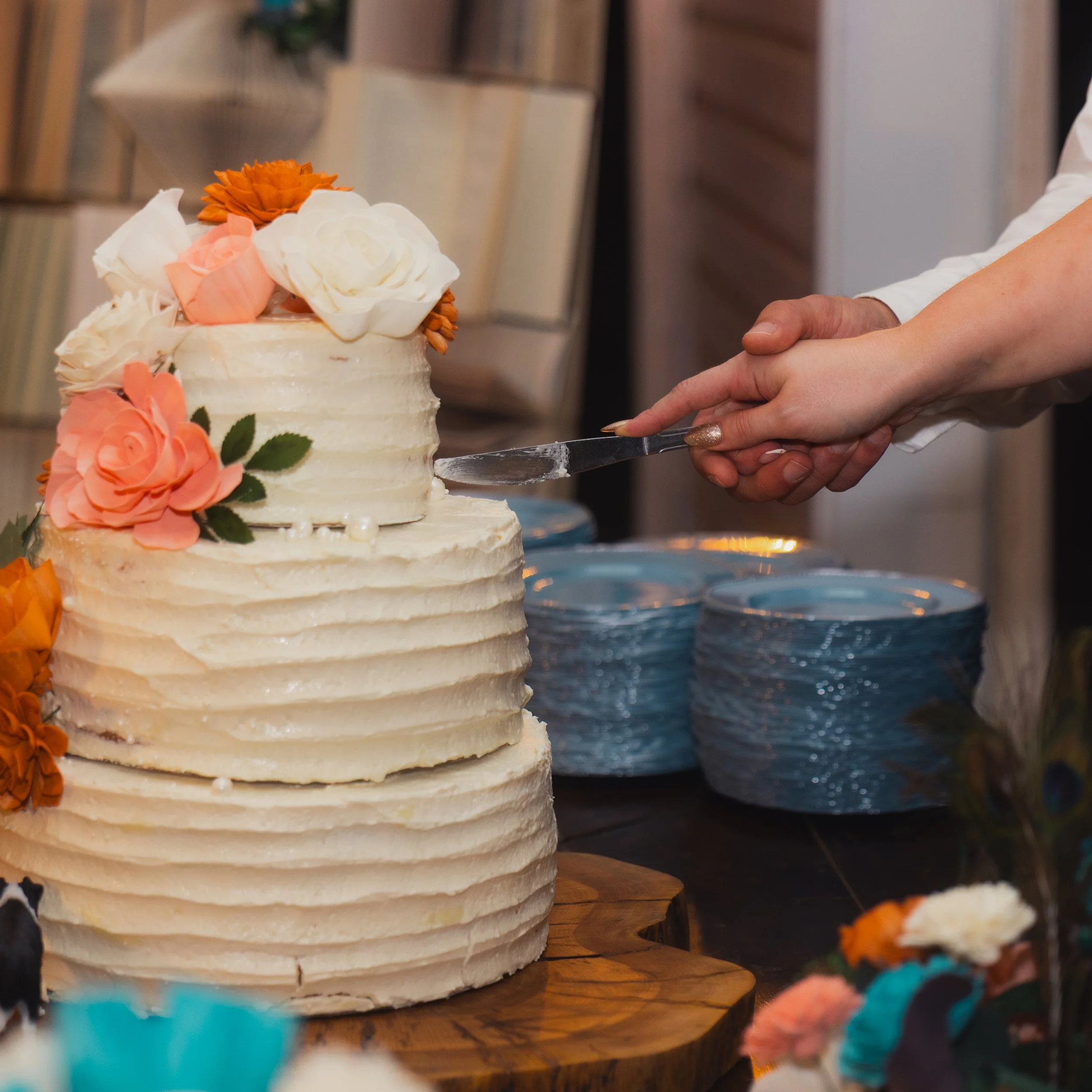 A person in white clothing is cutting a two-tiered wedding cake decorated with white, pink, and orange flowers.