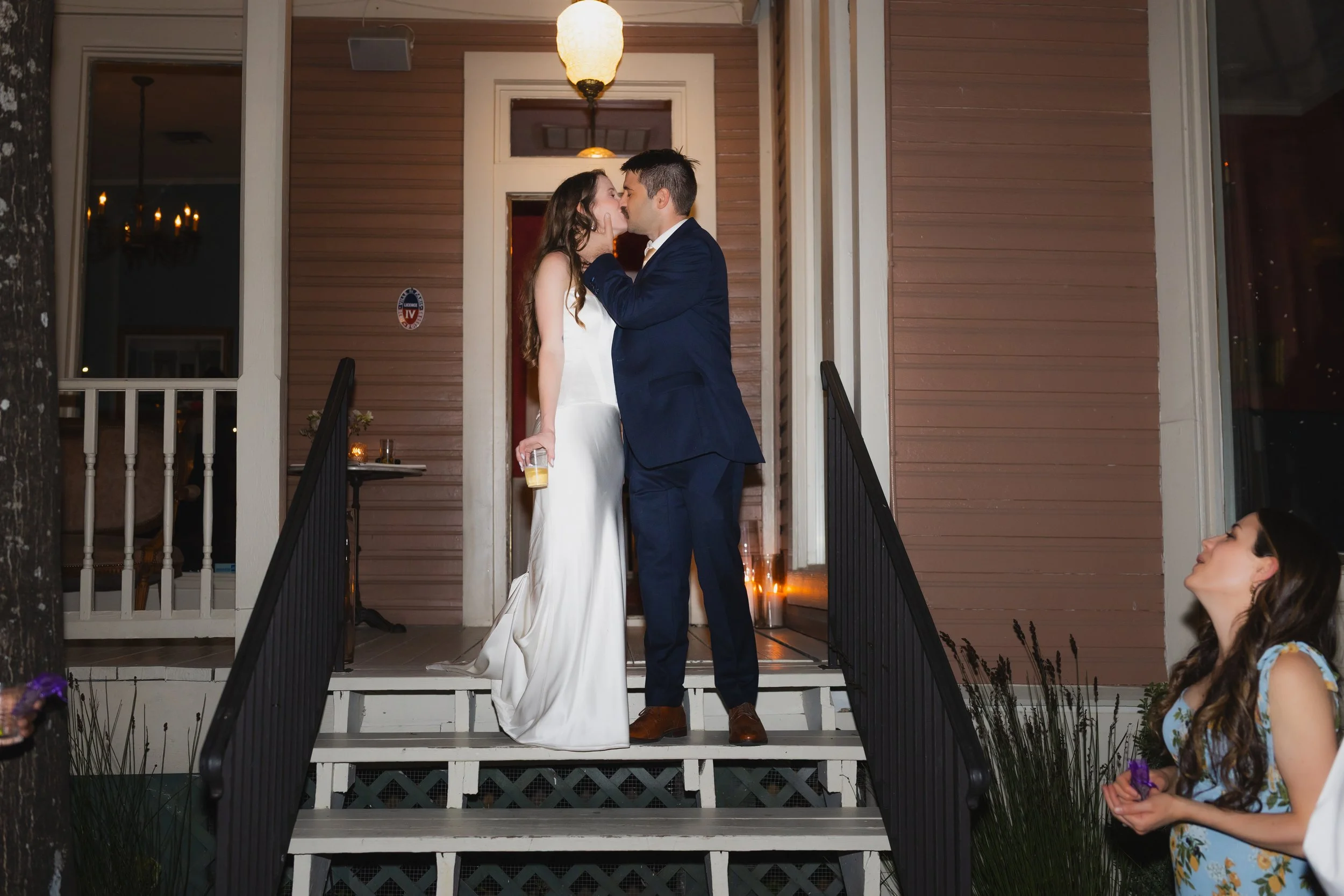 A couple in wedding attire kisses on the porch of a house, with a woman in a floral dress watching and smiling nearby.