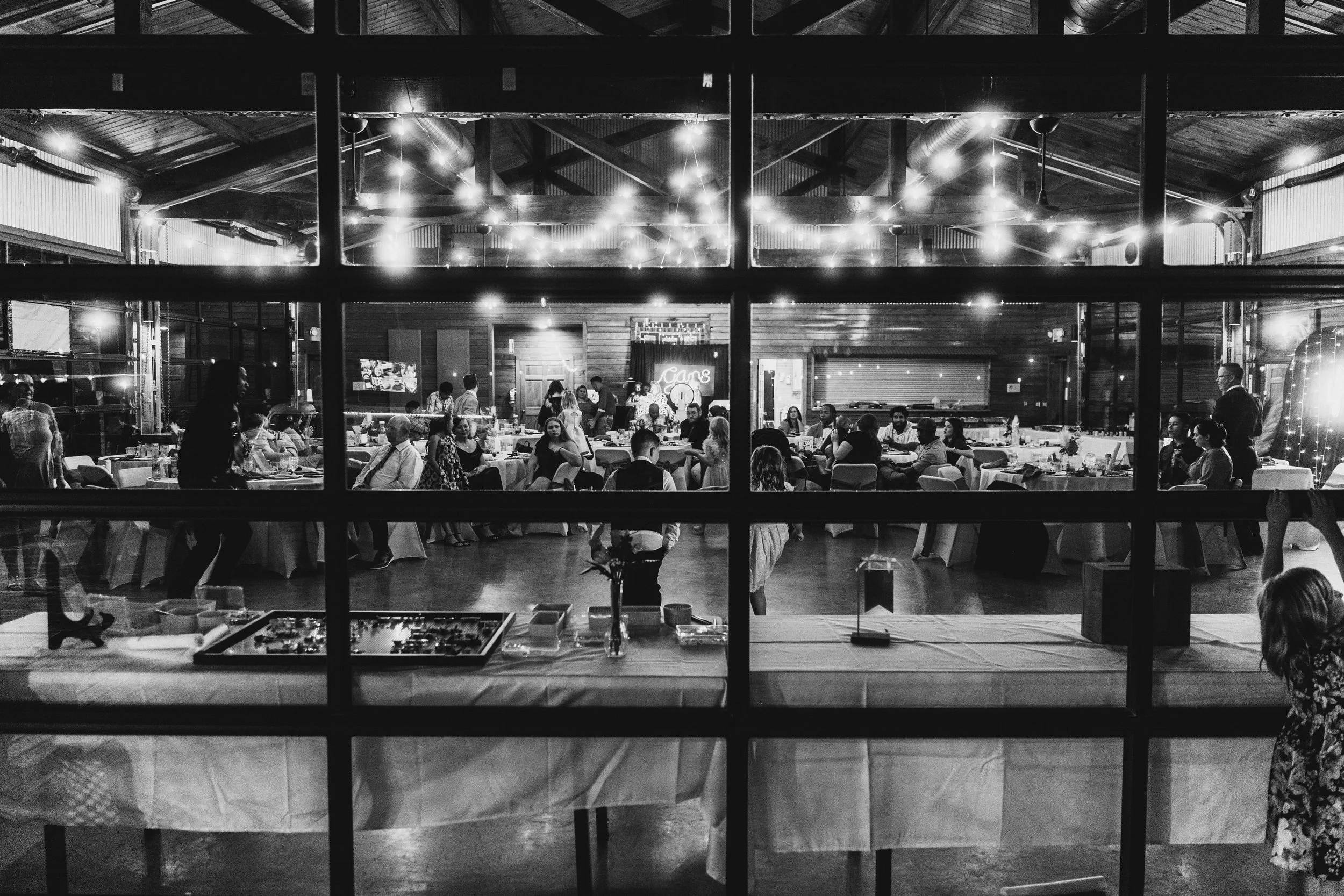 A black-and-white photo of a social gathering inside a rustic, wood-paneled event venue seen through a glass window divided into panes. There are multiple people seated at round tables decorated with flowers, with some standing and chatting. String l