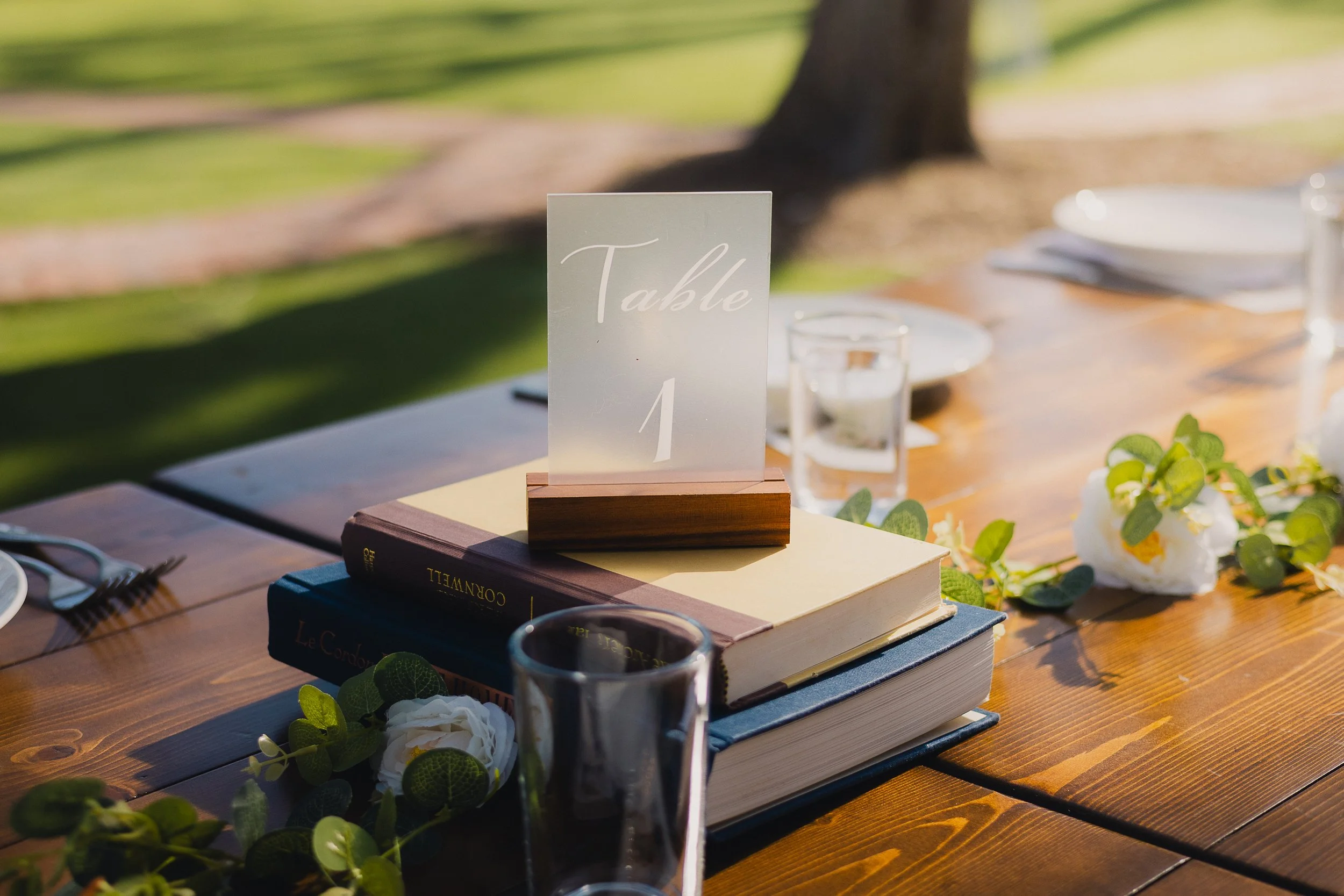 Wedding reception table with a sign reading 'Table 1' on top of books, surrounded by flower decorations and glassware, set outdoors on a wooden table.