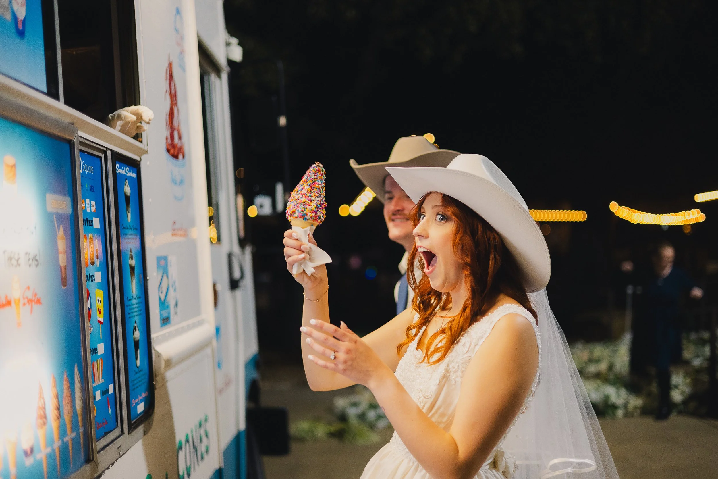A woman in a wedding dress and a large white hat with a veil, holding a colorful ice cream cone, smiling with her mouth open at an ice cream stand at night.