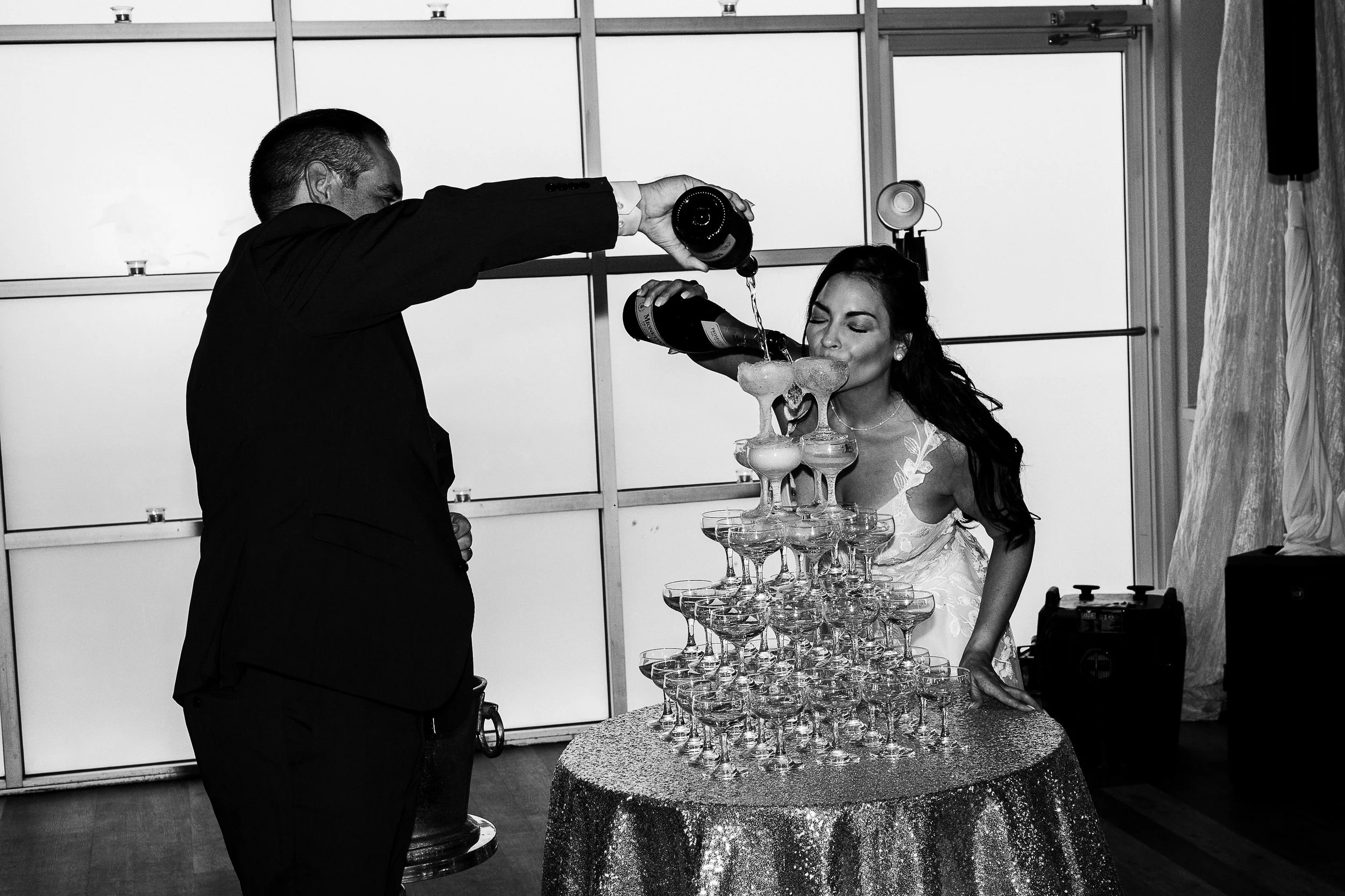 A man in a suit is pouring champagne into a tower of glasses held by a woman in a dress at a celebration or event.