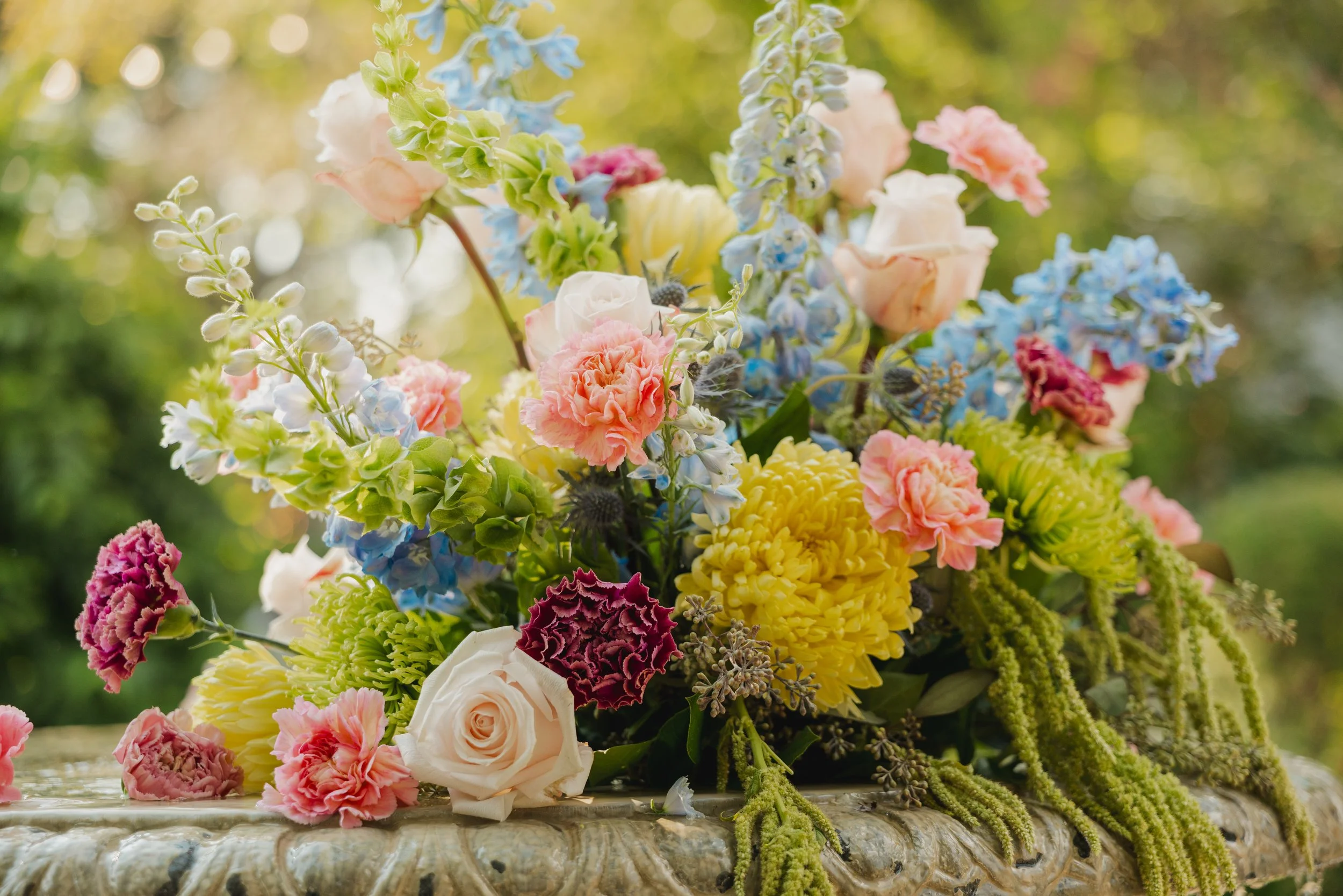 A colorful bouquet of various flowers including roses, carnations, delphiniums, and other blooms on a stone surface with a blurred green background.