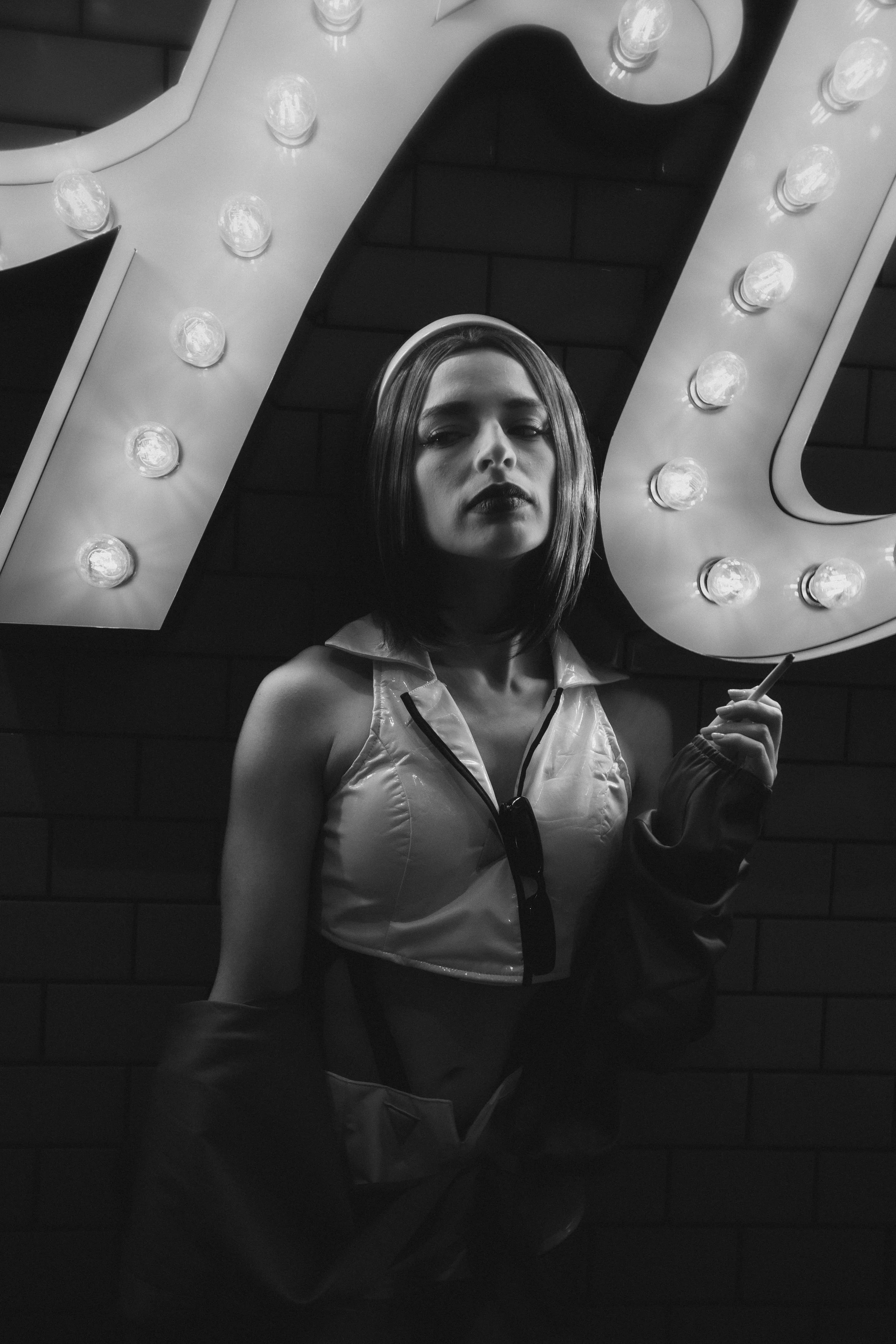 A young woman with shoulder-length dark hair and dark lipstick standing in front of a large illuminated sign, holding a cigarette, in black and white.