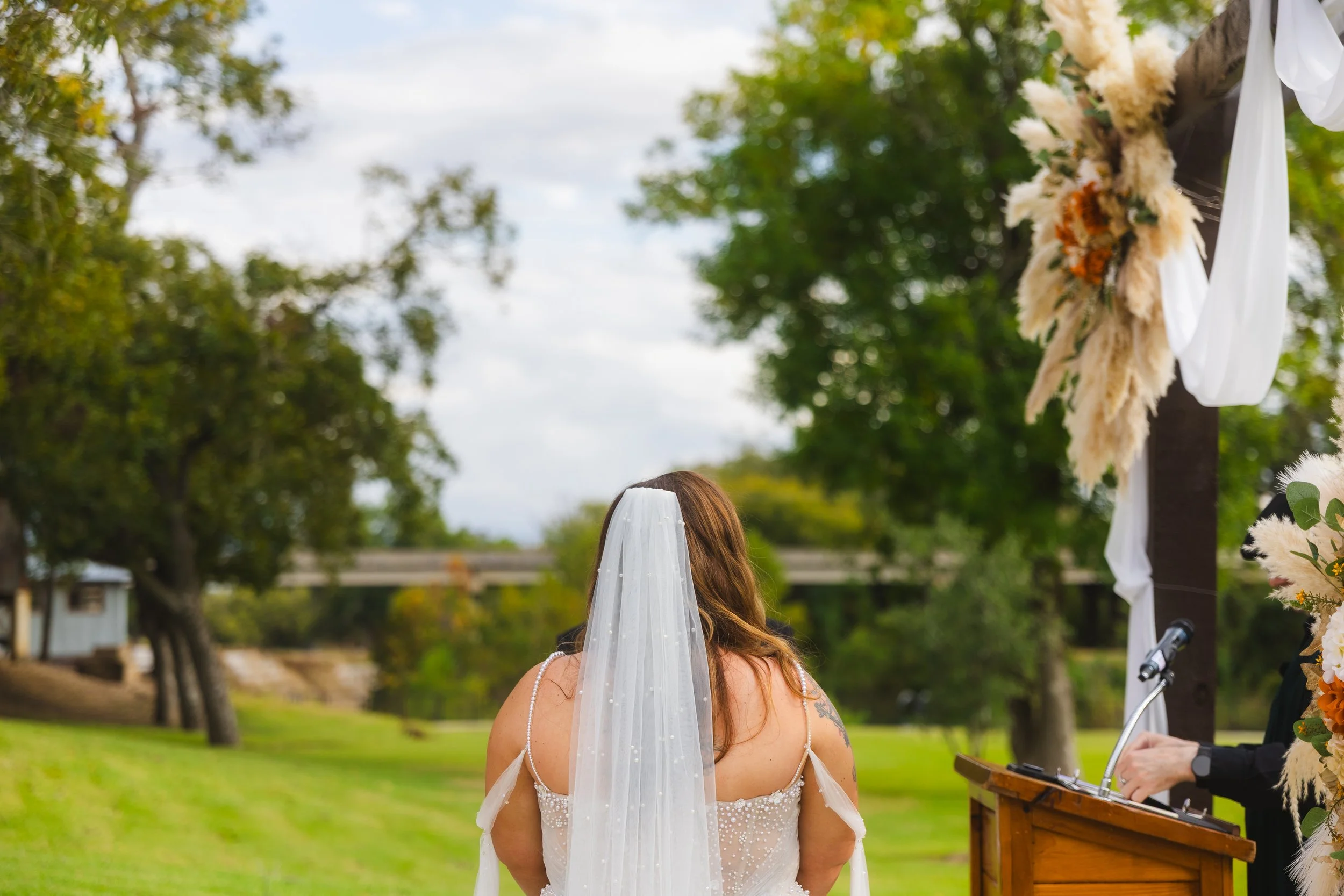 A bride with long brown hair wearing a white wedding dress and veil, standing outdoors at a wedding ceremony, facing an officiant at a wooden podium decorated with flowers and white fabric, surrounded by green trees and a cloudy sky.