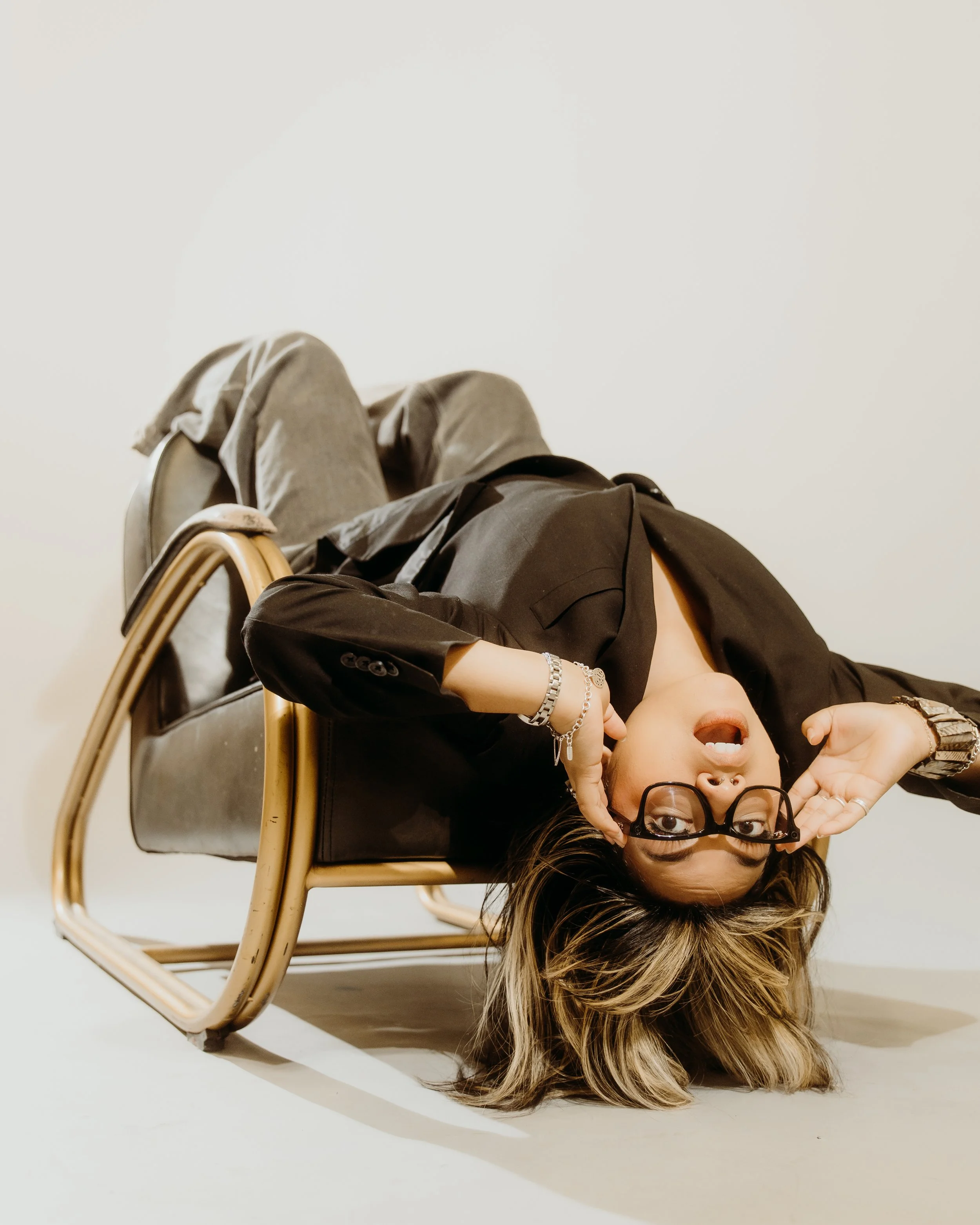 Woman lying on her back on a black and gold chair, with her head hanging upside down, holding glasses near her face, wearing a black blazer, jewelry, and gray pants.