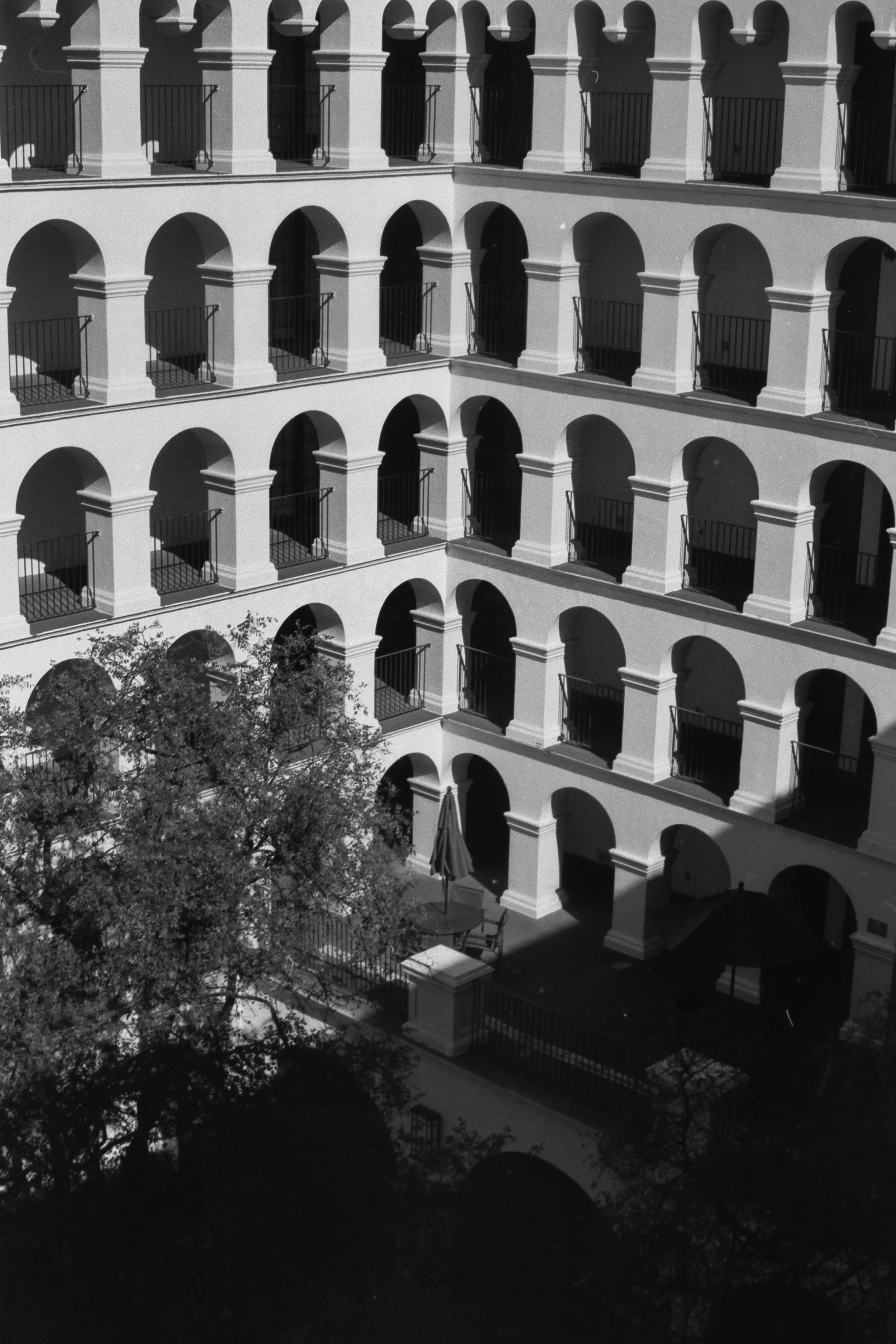Black and white photo of a multi-story building with multiple arched balconies and a courtyard with trees and umbrellas.