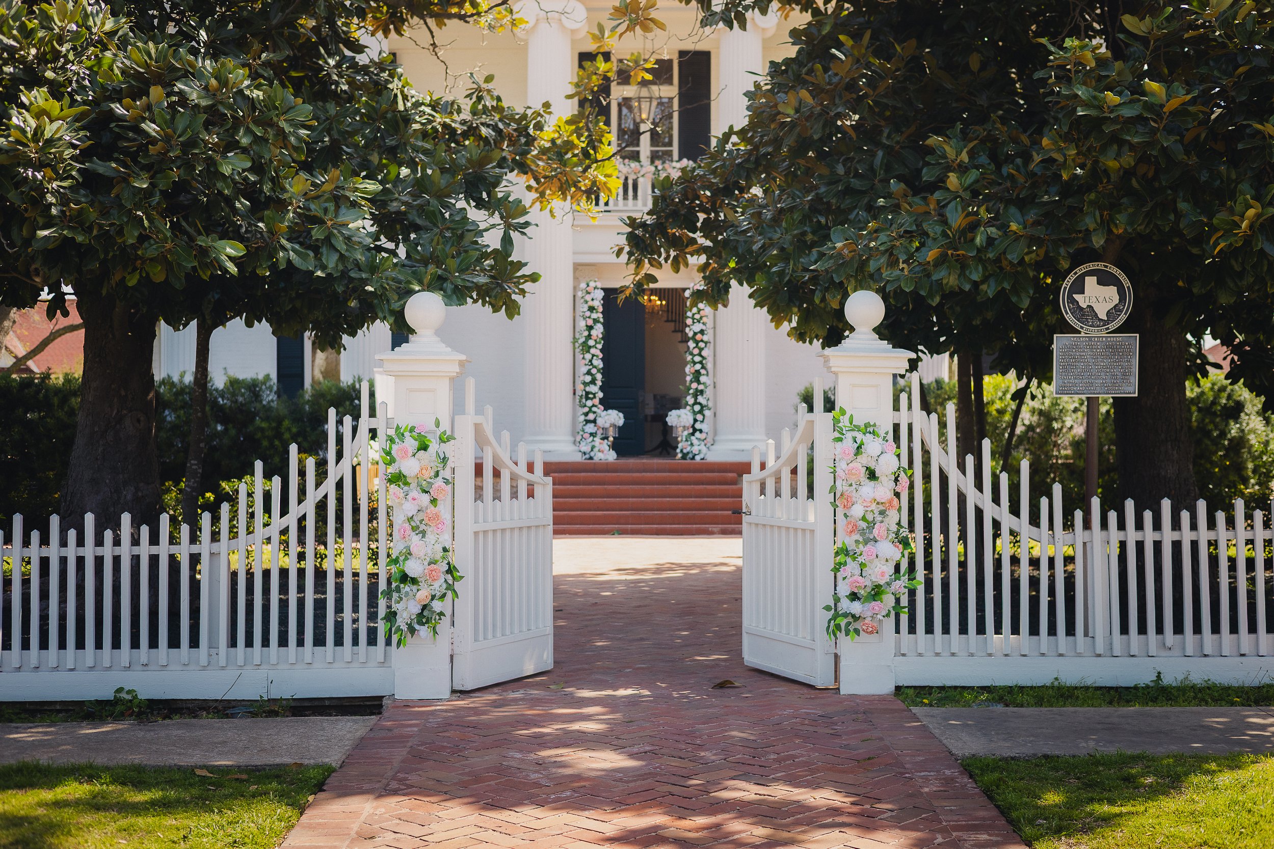 White picket fence gate decorated with pink and white flowers leading to a white house with pillars and steps, surrounded by green trees.