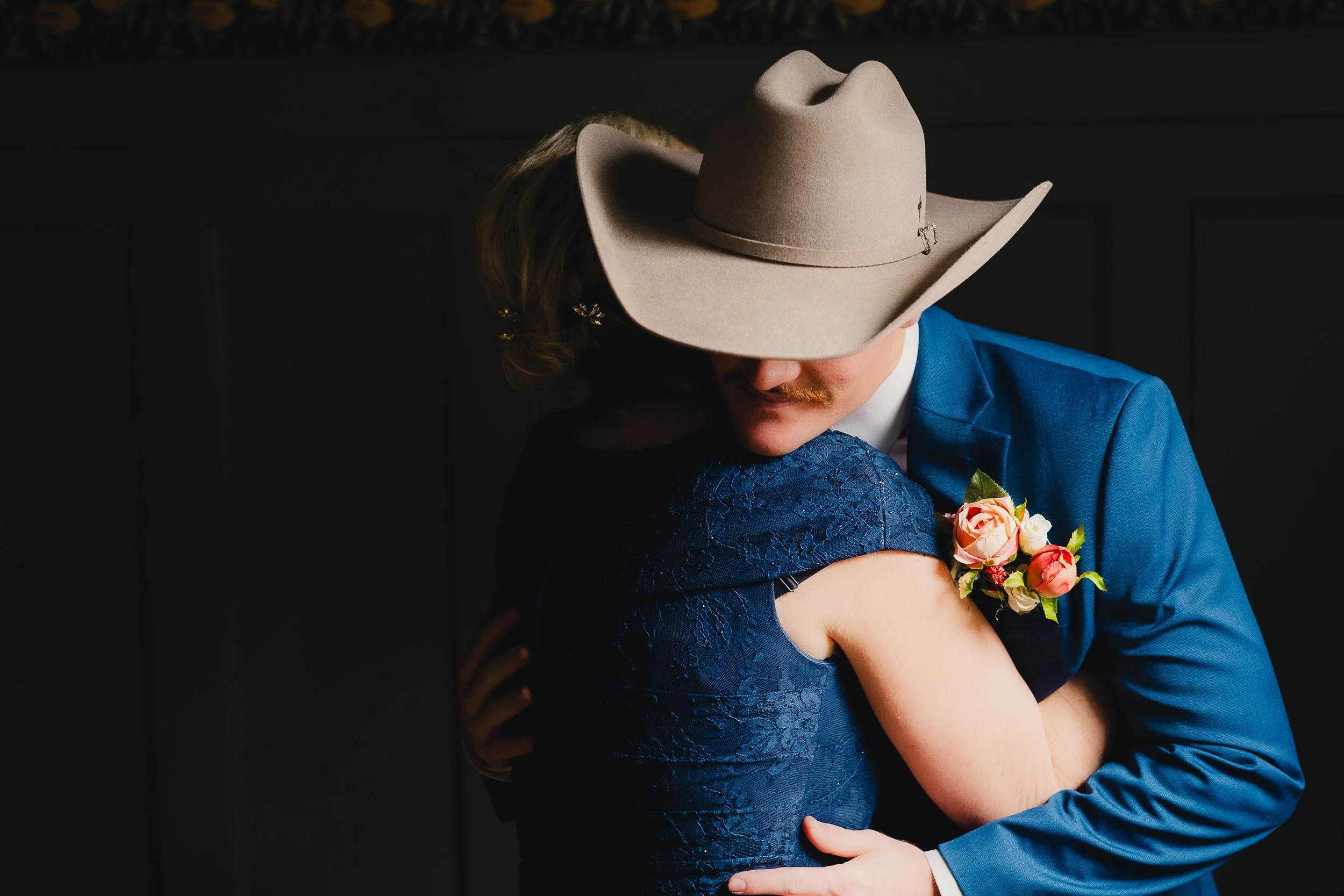 A man wearing a large beige cowboy hat and blue suit hugging a woman in a dark blue lace dress. The man has a flower boutonniere on his lapel, and they are sharing an embrace.