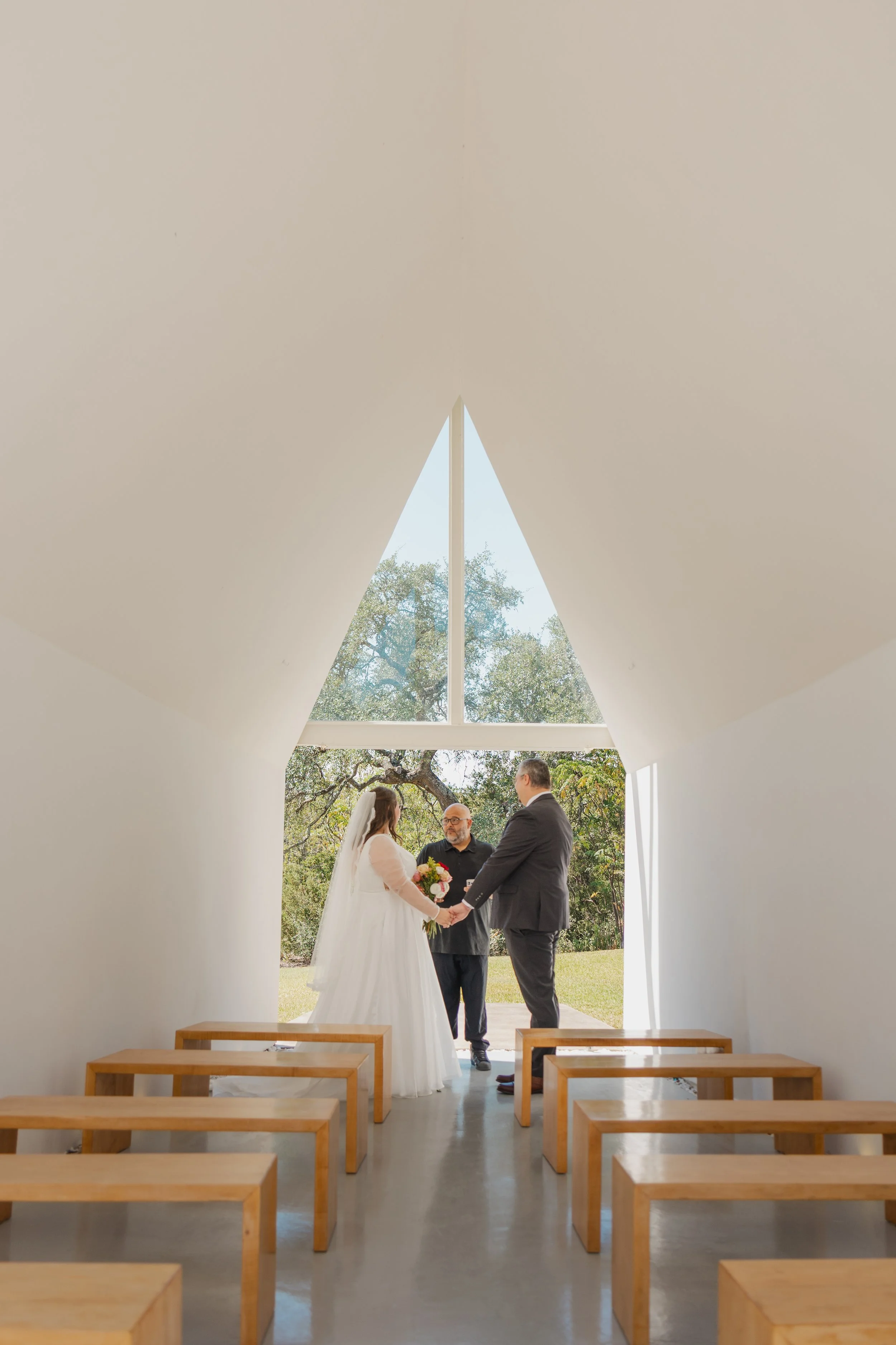 A wedding ceremony with a bride and groom holding hands and facing an officiant in a small chapel with large window and wooden benches.