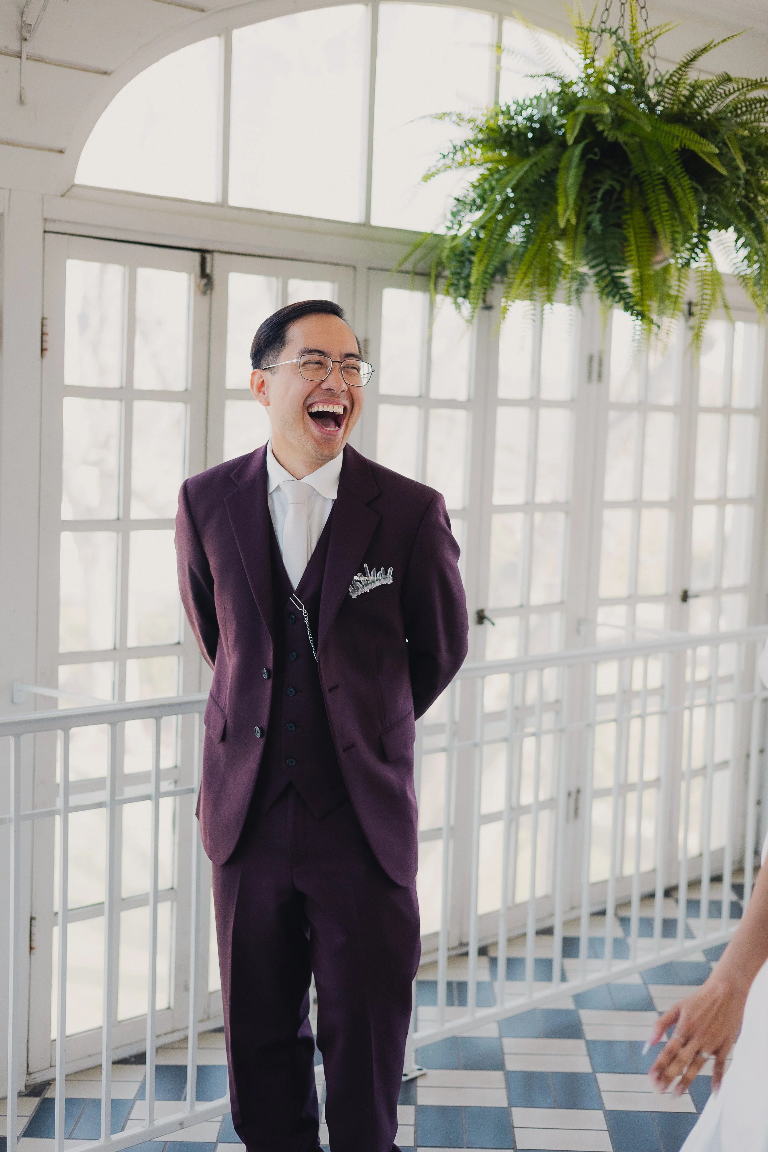 Man in a purple suit, white shirt, and glasses laughing in a bright indoor space with large windows and a hanging fern plant.