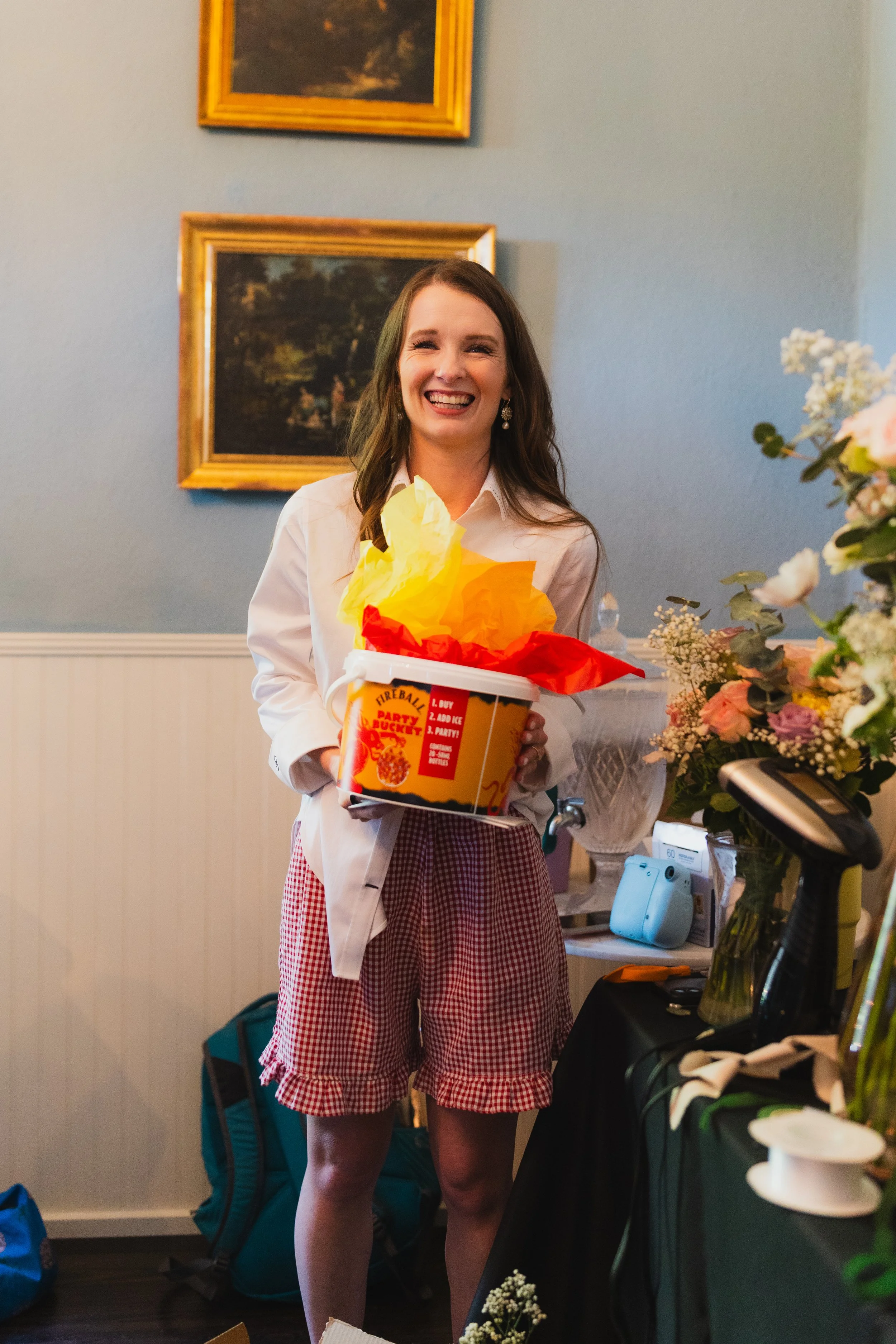 A young woman with long brown hair, wearing a white shirt and red checkered shorts, is smiling while holding a bucket of popcorn with yellow, red, and orange tissue paper inside. She is standing indoors next to a table with flowers and various items.