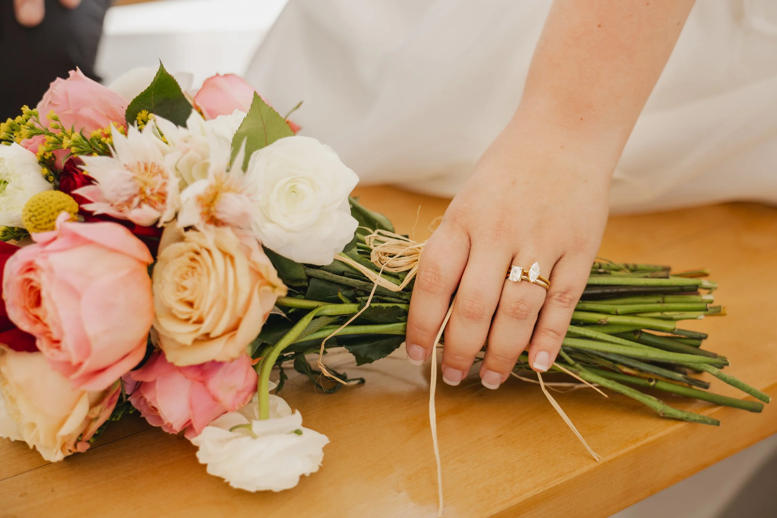 A person’s hand adjusting a bouquet of pink, white, and cream flowers on a wooden surface, wearing a ring with a large diamond.