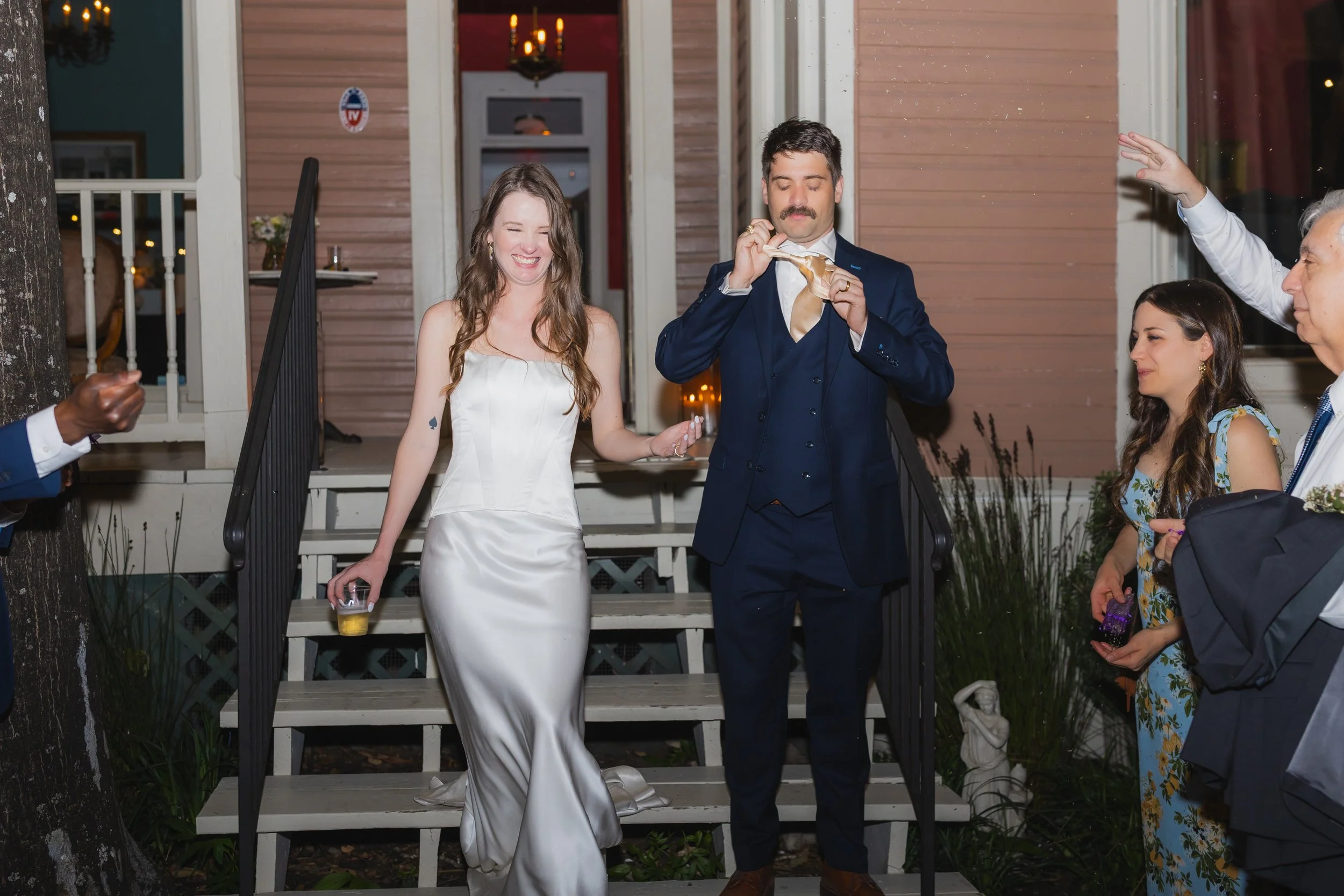 A wedding celebration with a bride in a white satin gown and a groom in a navy suit, standing on stairs outside a house. The bride is smiling and holding a drink, while the groom is removing his tie. Guests are around them, some clapping and others w