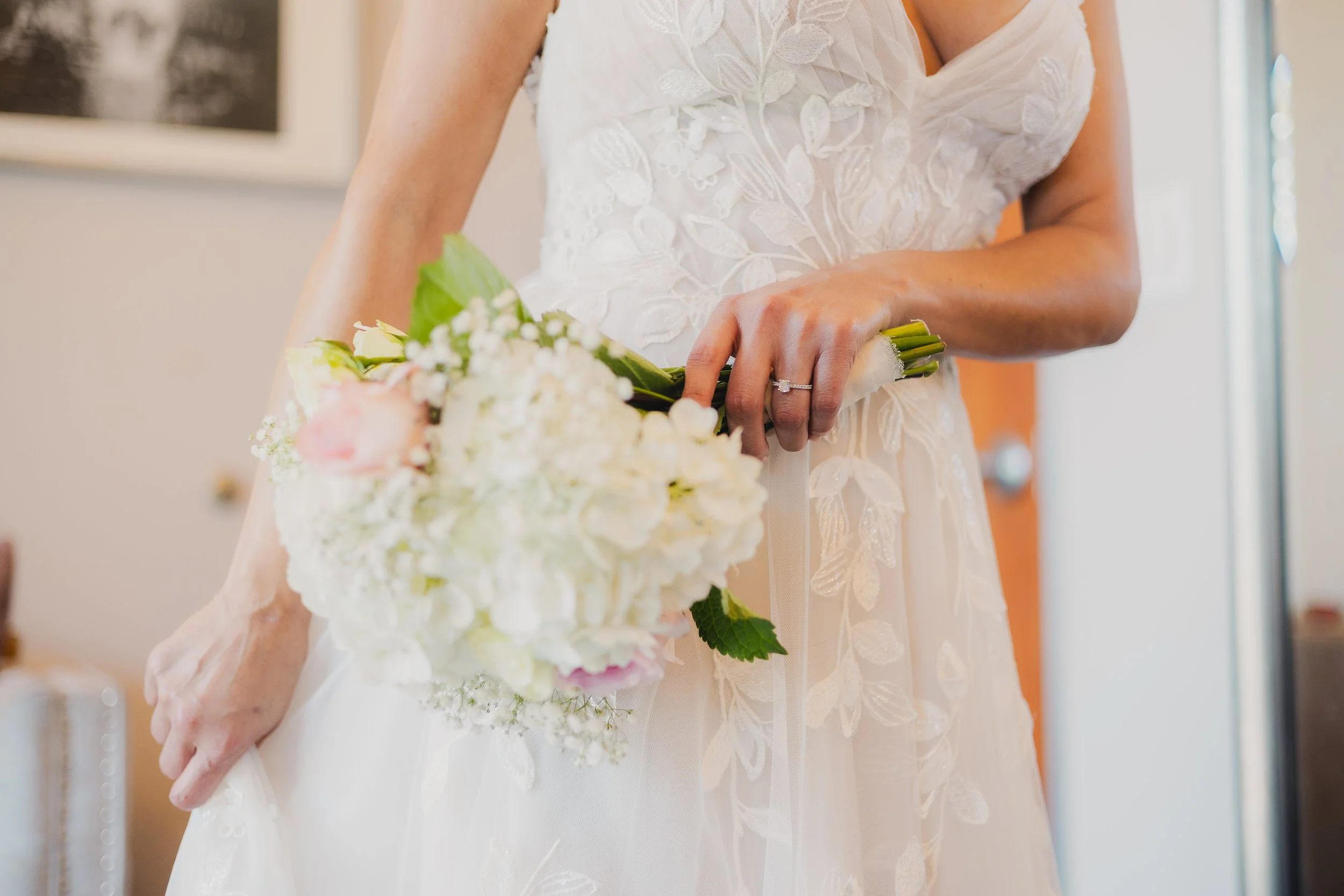 Close-up of a bride holding a bouquet of white and pink flowers, wearing a white lace wedding dress, with her wedding ring visible on her finger.