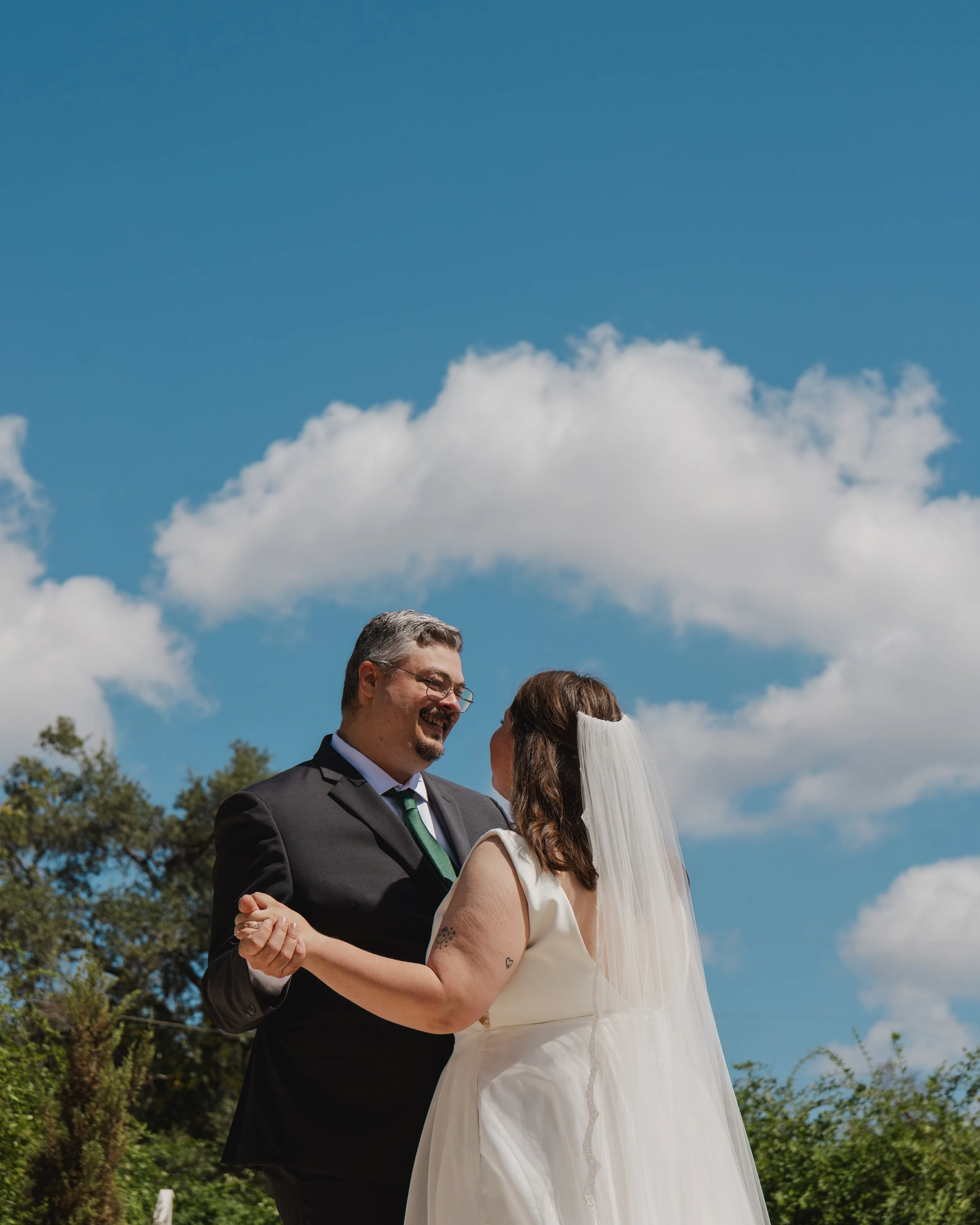 A couple in wedding attire dancing outdoors under a cloudy blue sky.