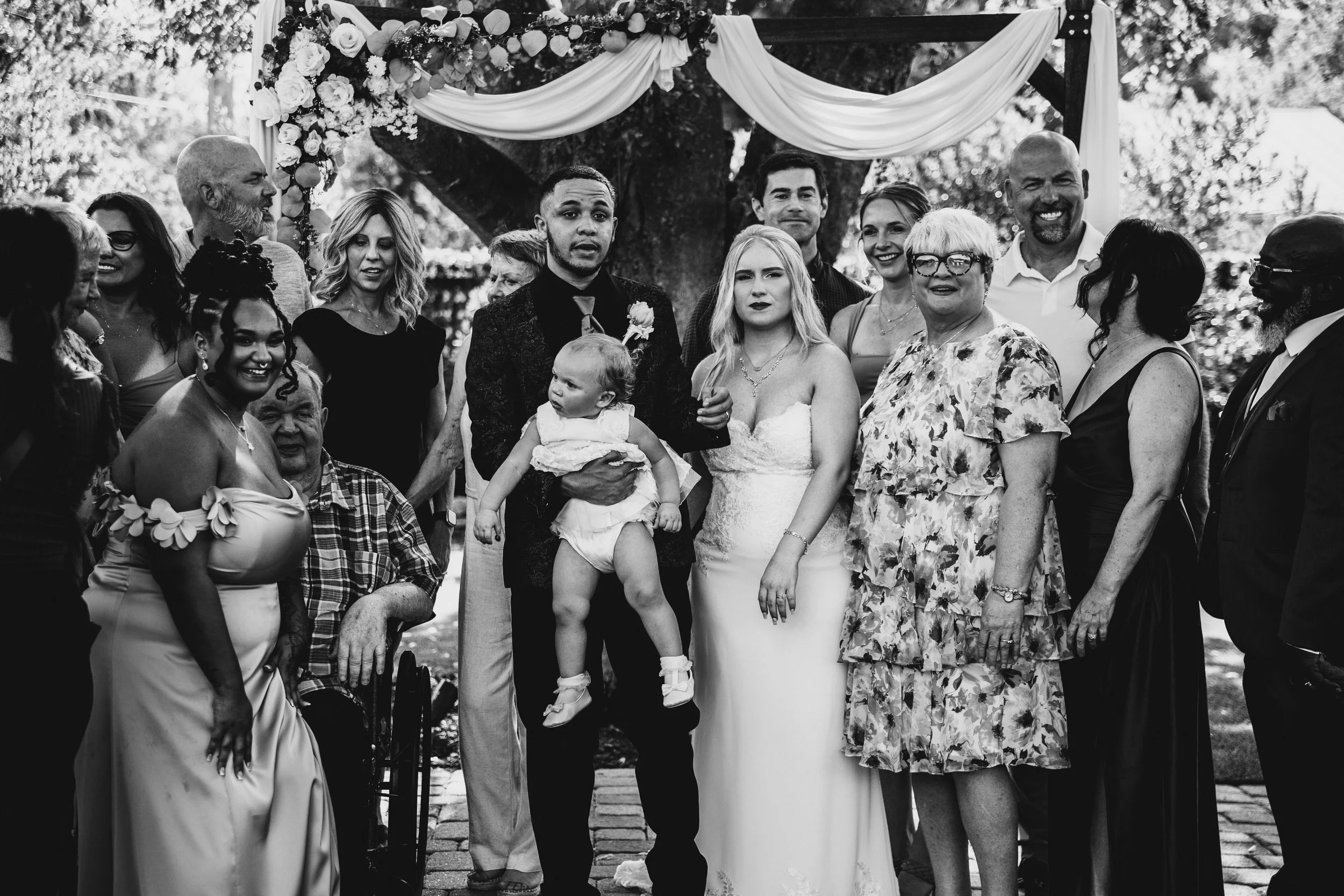 A black and white photo of a group of people gathered outdoors at a wedding, standing under a decorated arch with flowers and drapes, including the bride and groom at the center surrounded by family and friends.