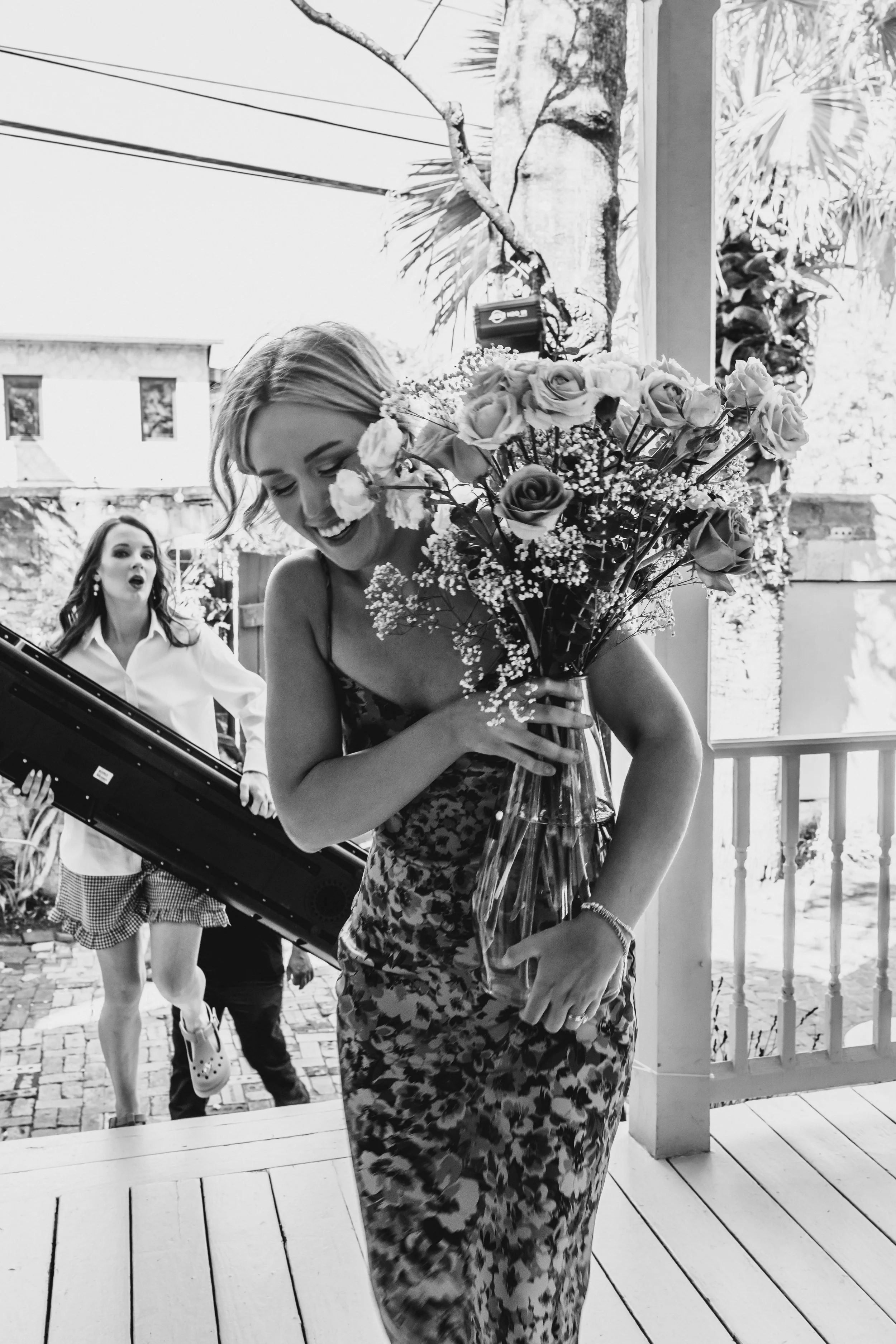 A woman holding a large bouquet of roses and baby's breath, smiling, with another woman in the background surprised, outdoors on a porch with trees and a building visible.