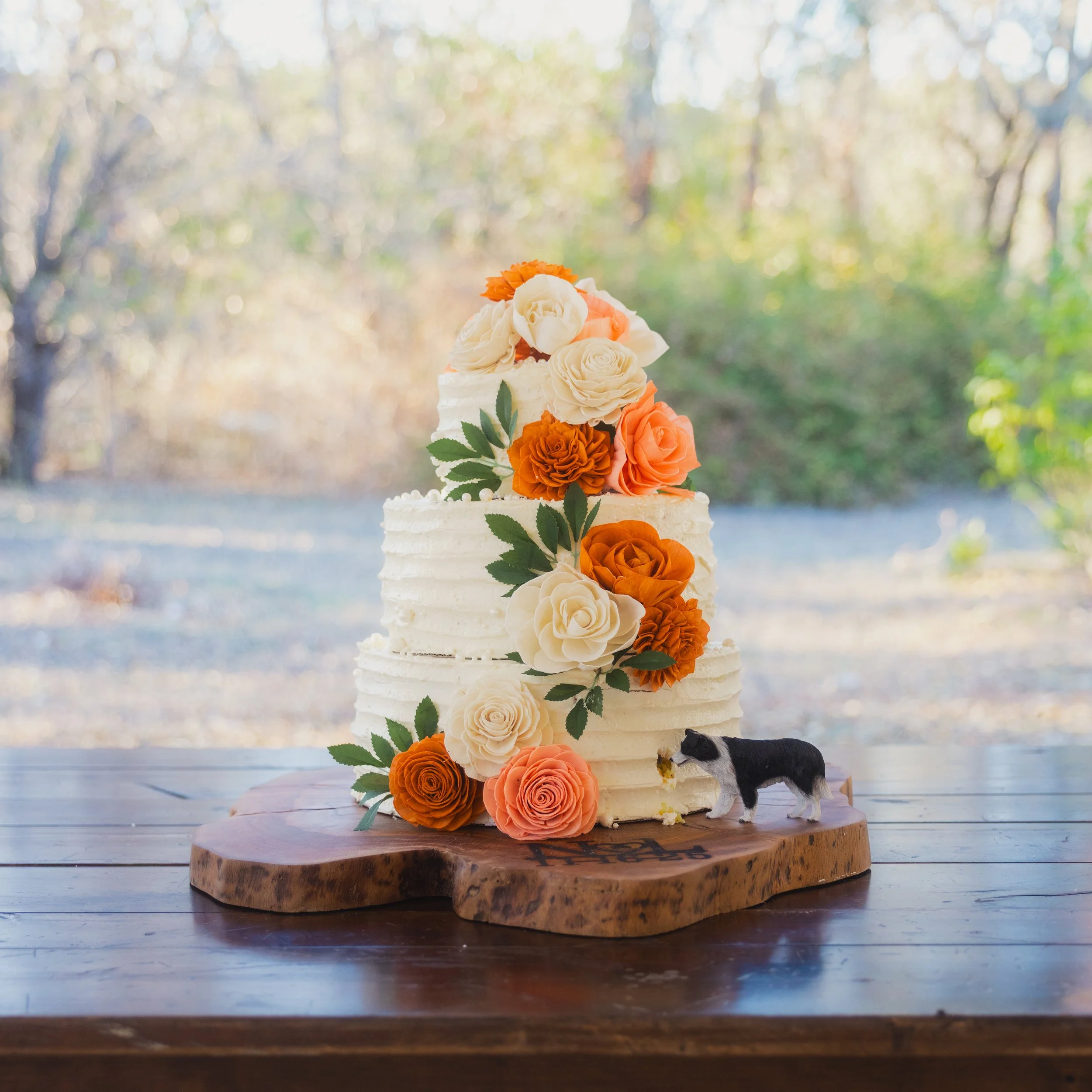 A three-tier white wedding cake decorated with orange and white roses and green leaves, placed on a wooden slab on a wooden table in an outdoor setting with trees in the background.