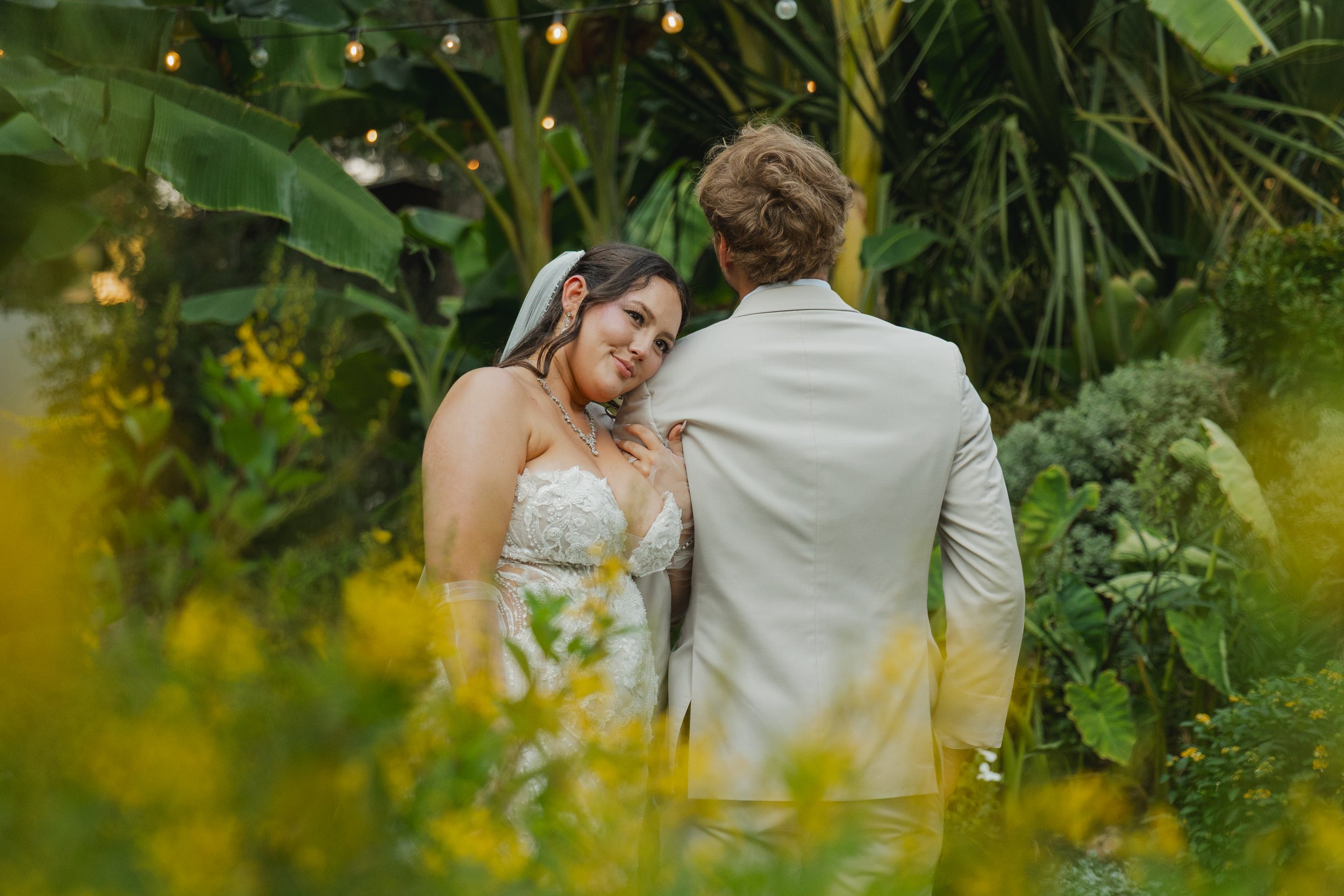 Bride and groom standing close together in a lush, green garden, with the bride leaning on the groom's shoulder, smiling softly, wearing a white wedding dress, a necklace, and a veil.