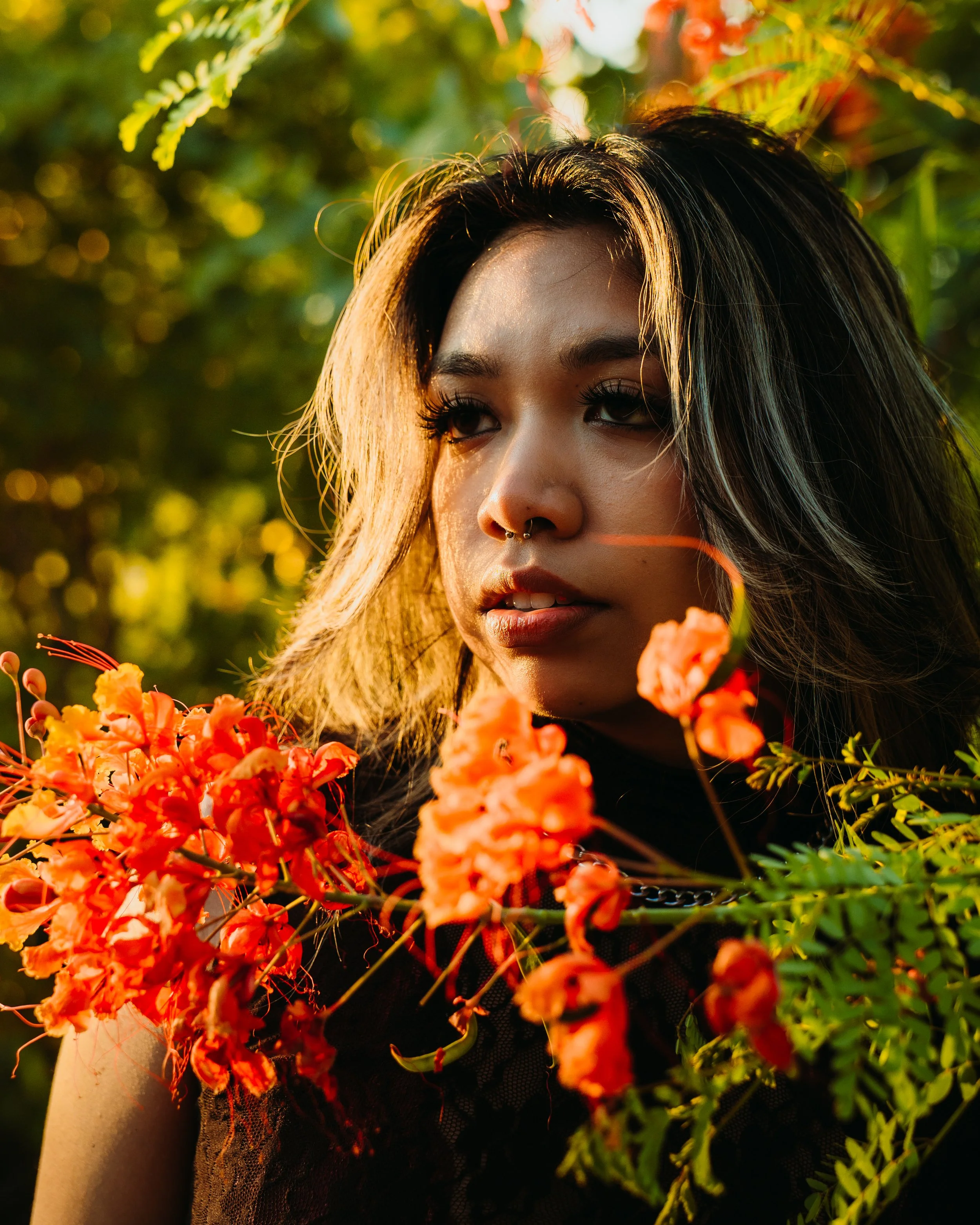 A woman with dark hair and septum piercing surrounded by orange flowers outdoors during golden hour.