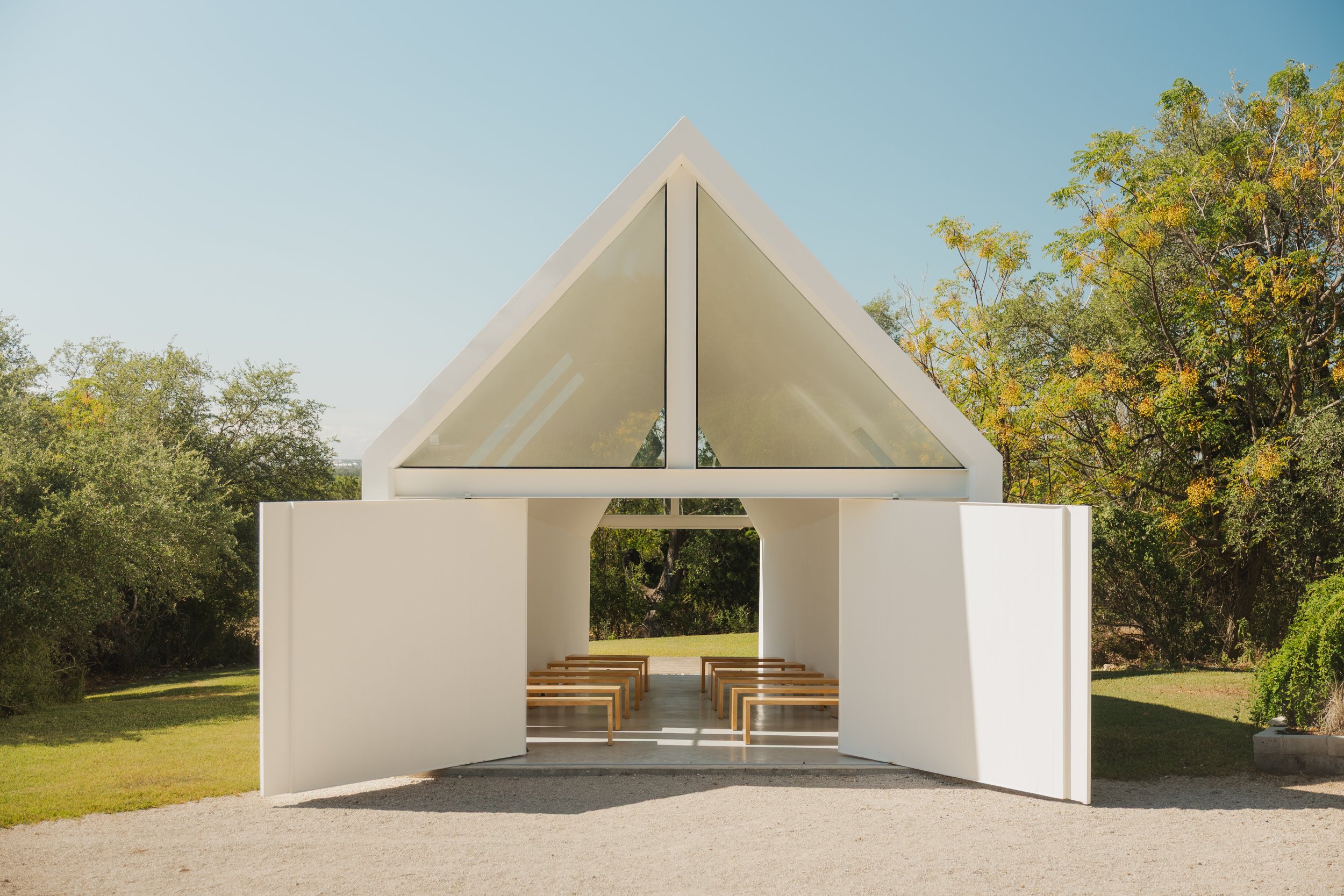 Modern white chapel with large glass windows and open doors, surrounded by greenery and trees under a clear blue sky.