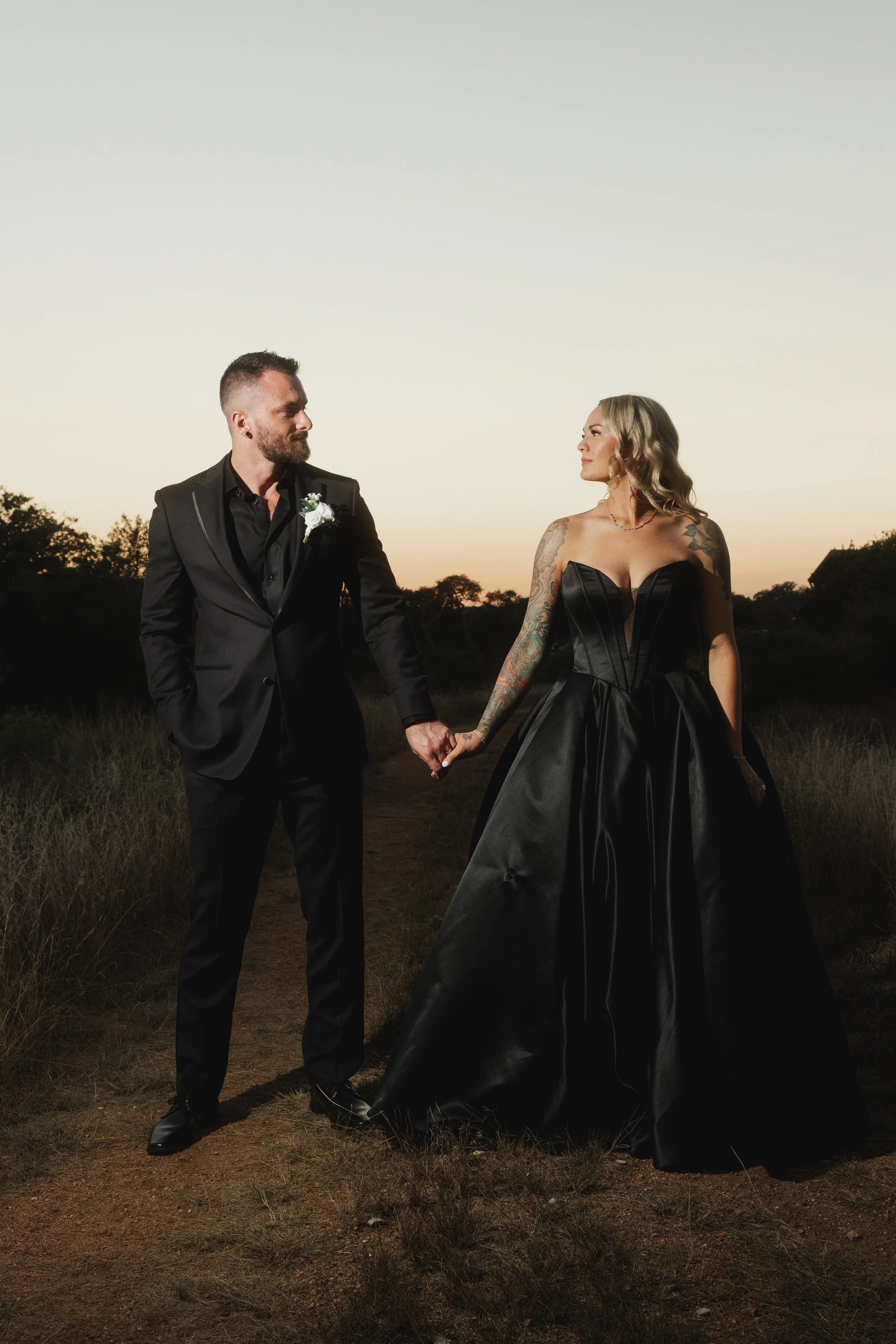 A couple holding hands outdoors at sunset, dressed in formal black attire, with the woman wearing a strapless black gown and the man in a black suit.