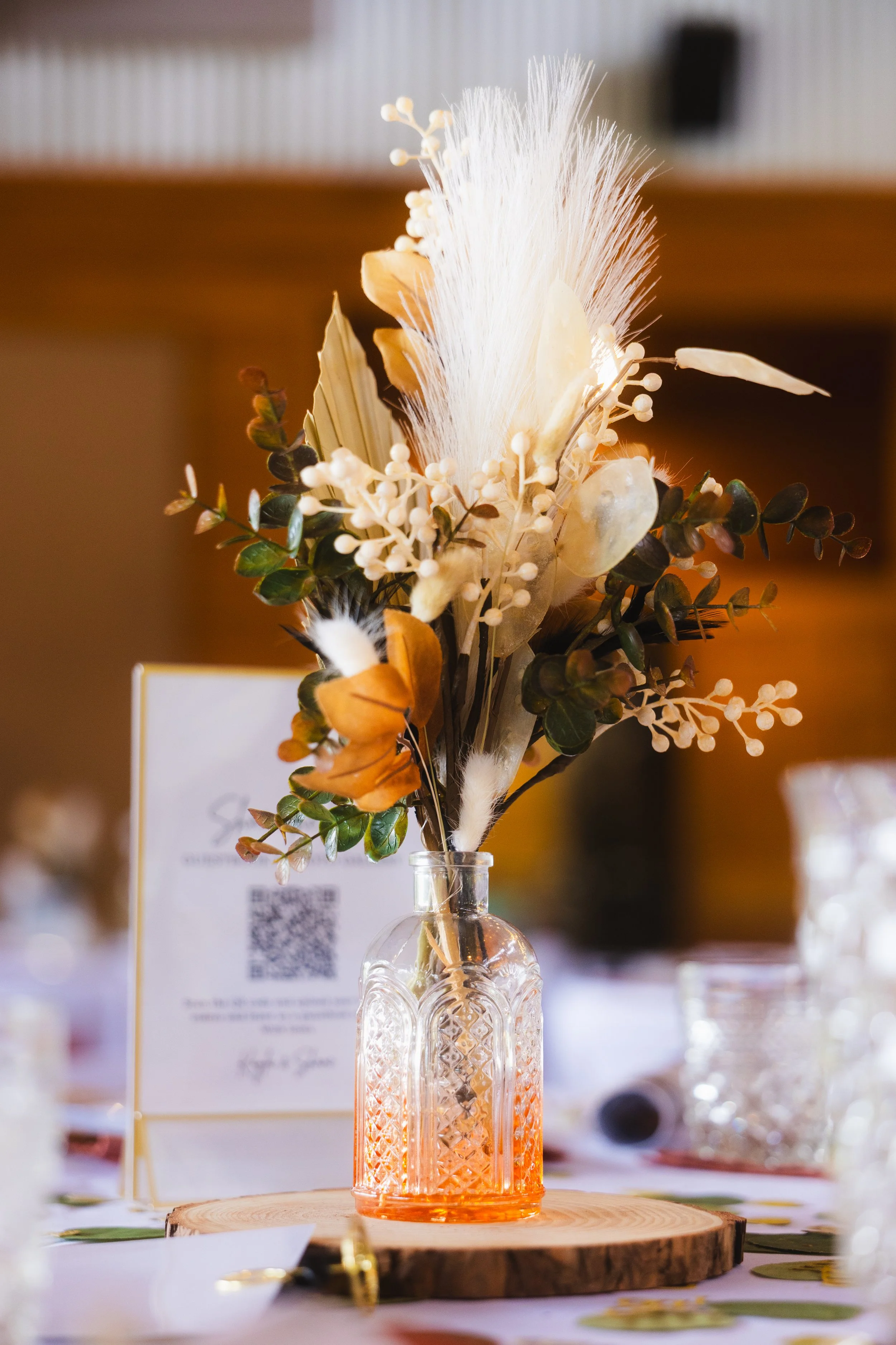 A decorative floral arrangement in an orange-tinted glass vase on a wooden slice, featuring dried flowers, leaves, and feathers, placed on a table with a blurred background.