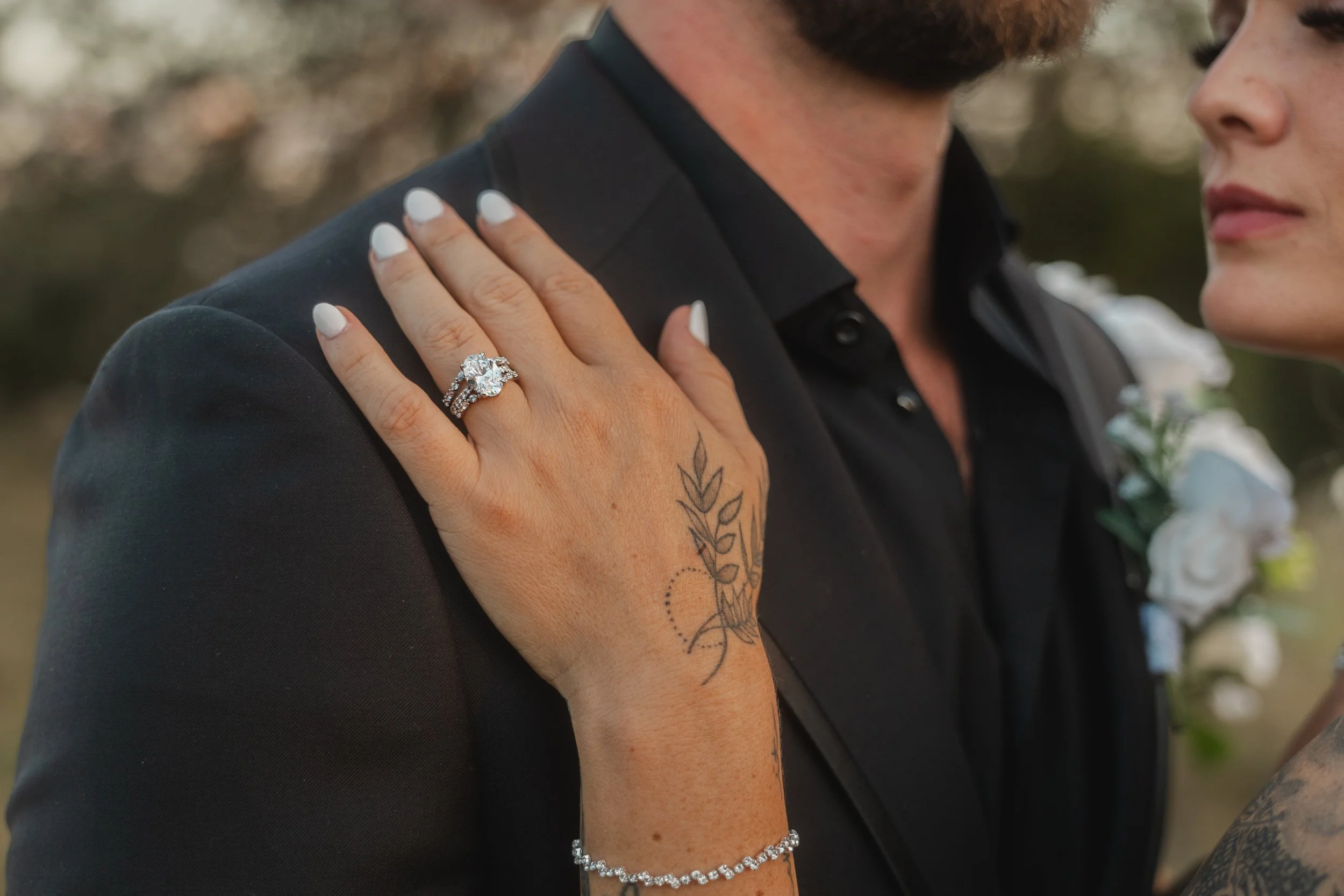 Close-up of a woman’s hand with a wedding ring, placed on a man’s shoulder, with a tattoo visible on her wrist and a bracelet. The man is wearing a black jacket with a boutonniere featuring white flowers, and their faces are partially visible against