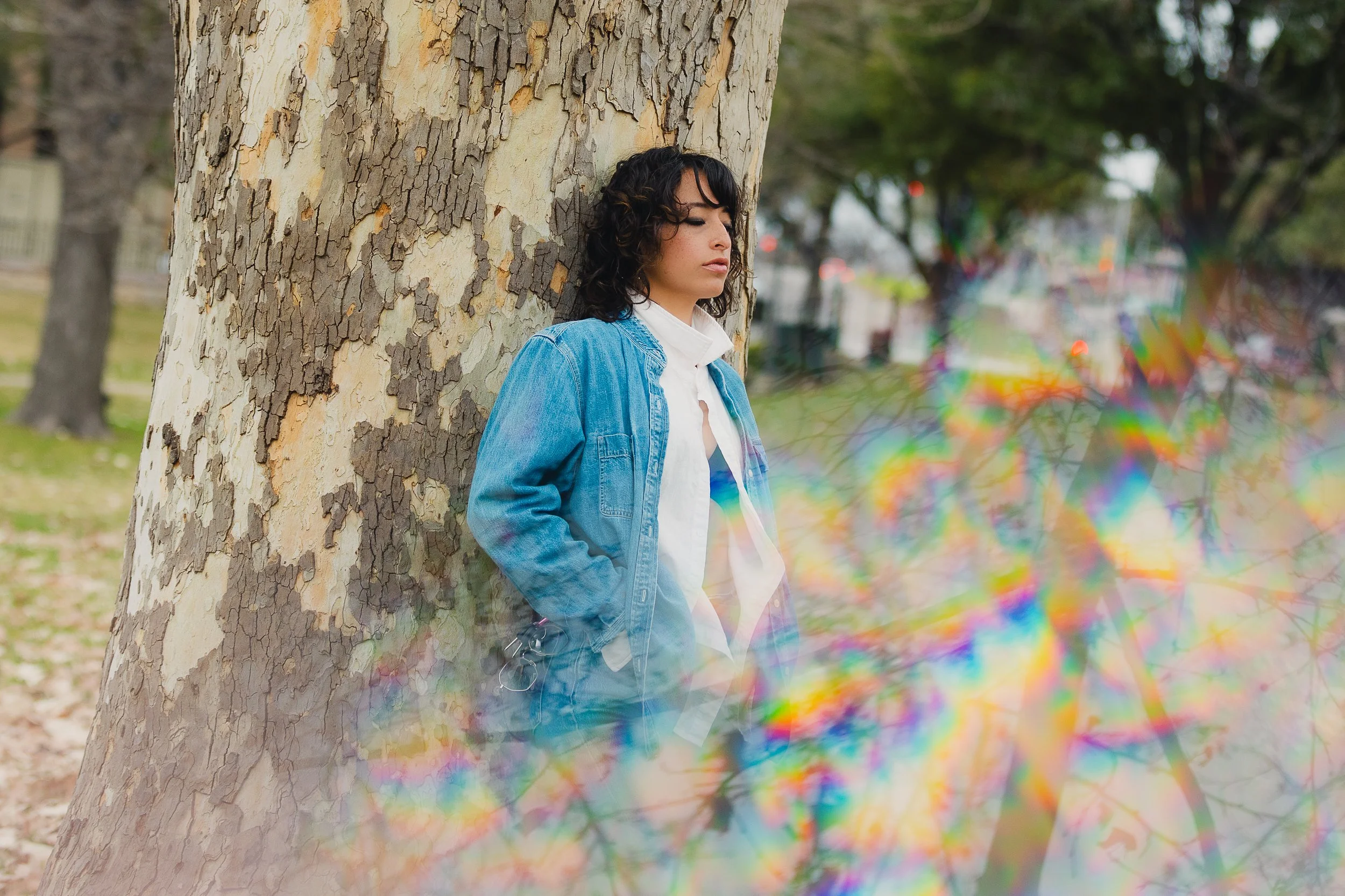 A woman with dark curly hair wearing a white shirt and blue denim jacket leaning against a large tree in a park, with colorful rainbow reflections in the foreground.