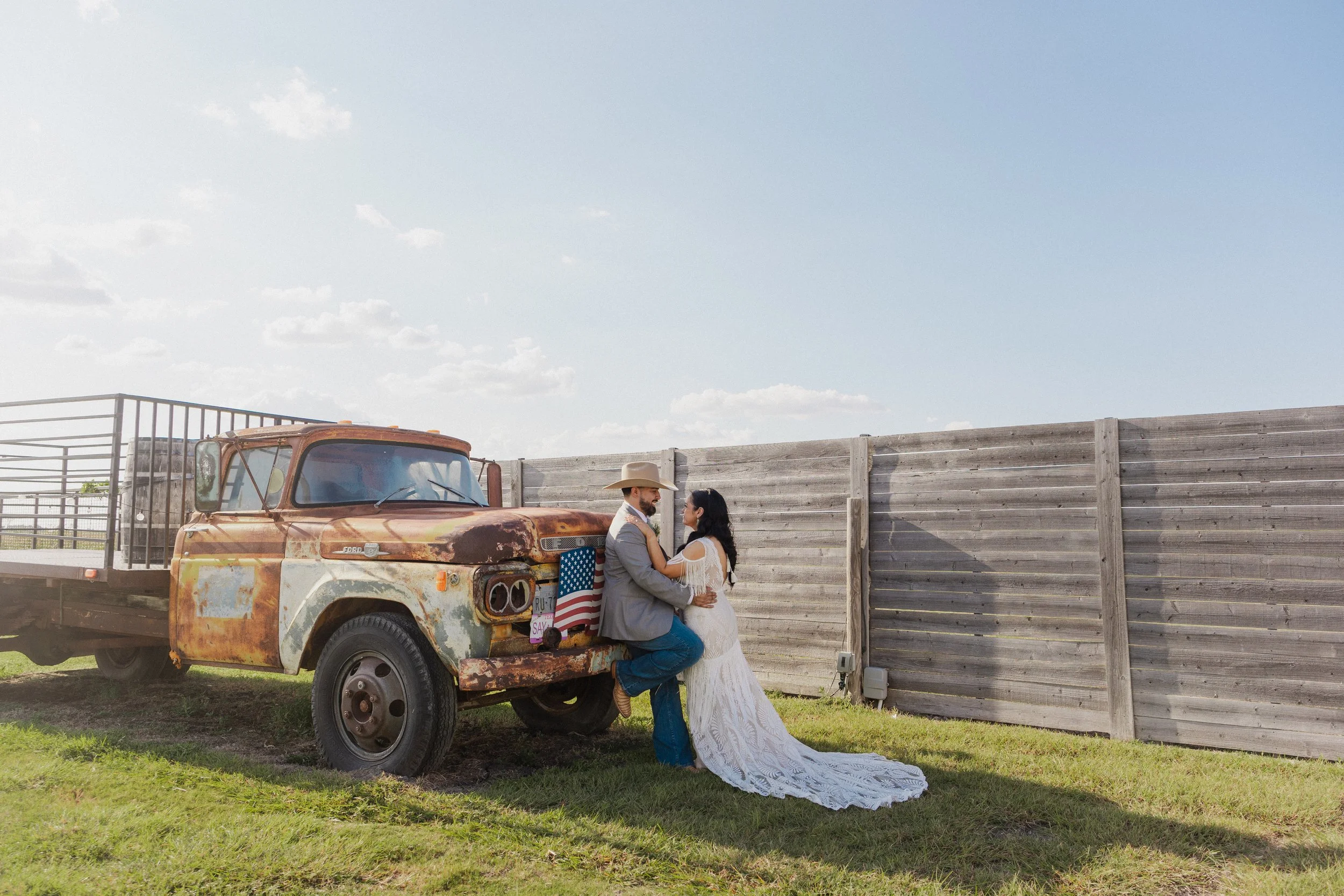 A couple dressed in wedding attire is sitting on the front of a vintage, rusty pickup truck with an American flag decoration. They are outdoors near a wooden fence and a grassy area, with a large open sky above.