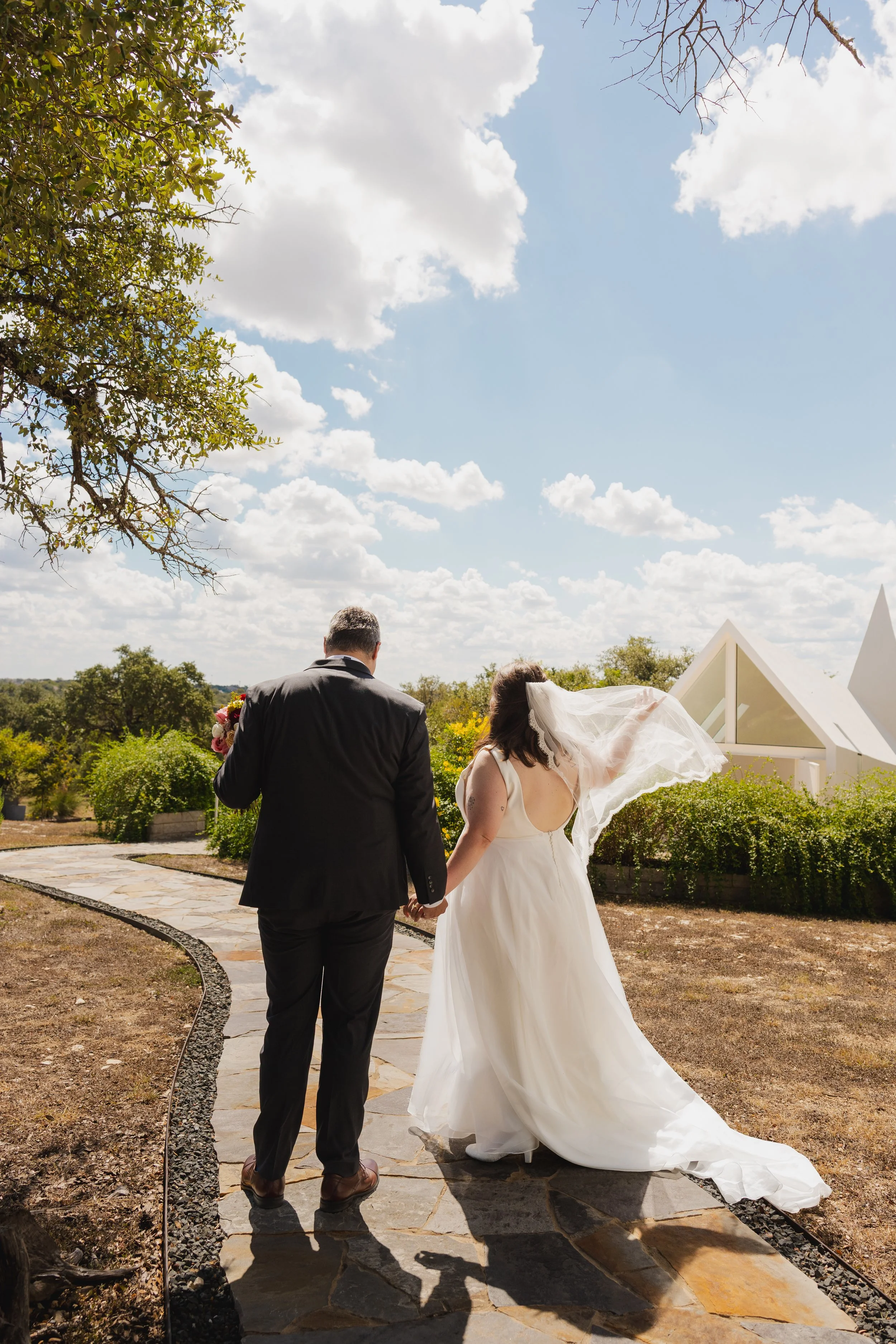 A couple, dressed in wedding attire, walking hand in hand outdoors under a partly cloudy sky, with trees and a small building in the background.