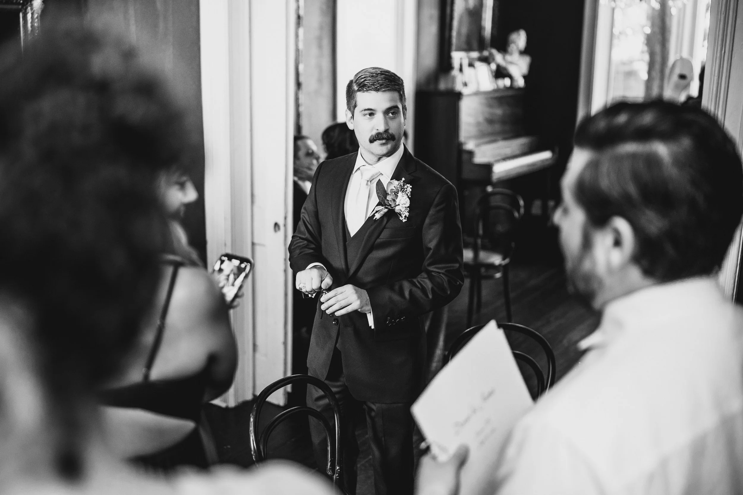 Black and white photo of a groom in a suit with a flower on his lapel, standing and talking to guests at a wedding reception inside a room with wood floors and large windows.
