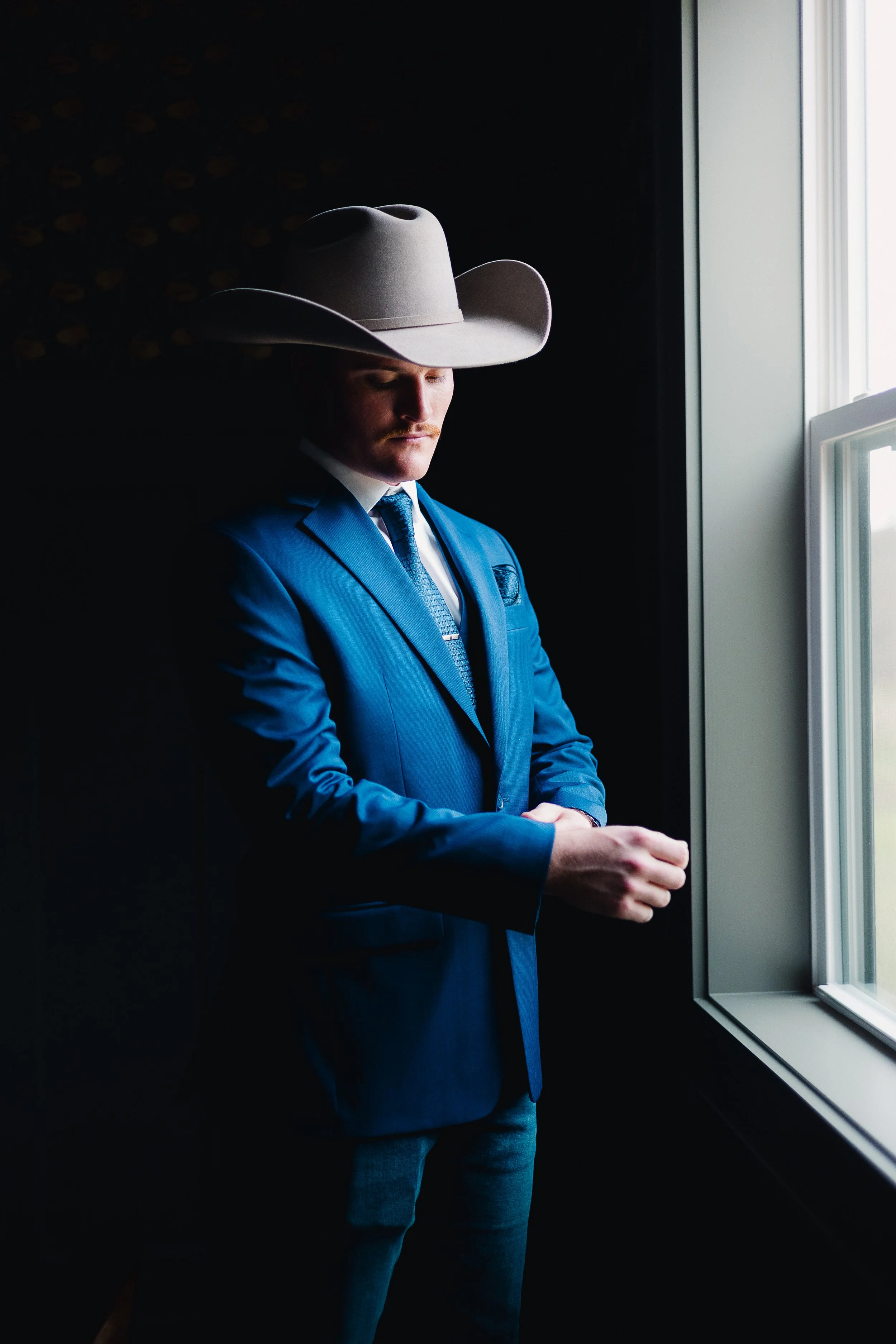 A man dressed in a blue suit and wearing a large cowboy hat, standing by a window, looking down with a contemplative expression.