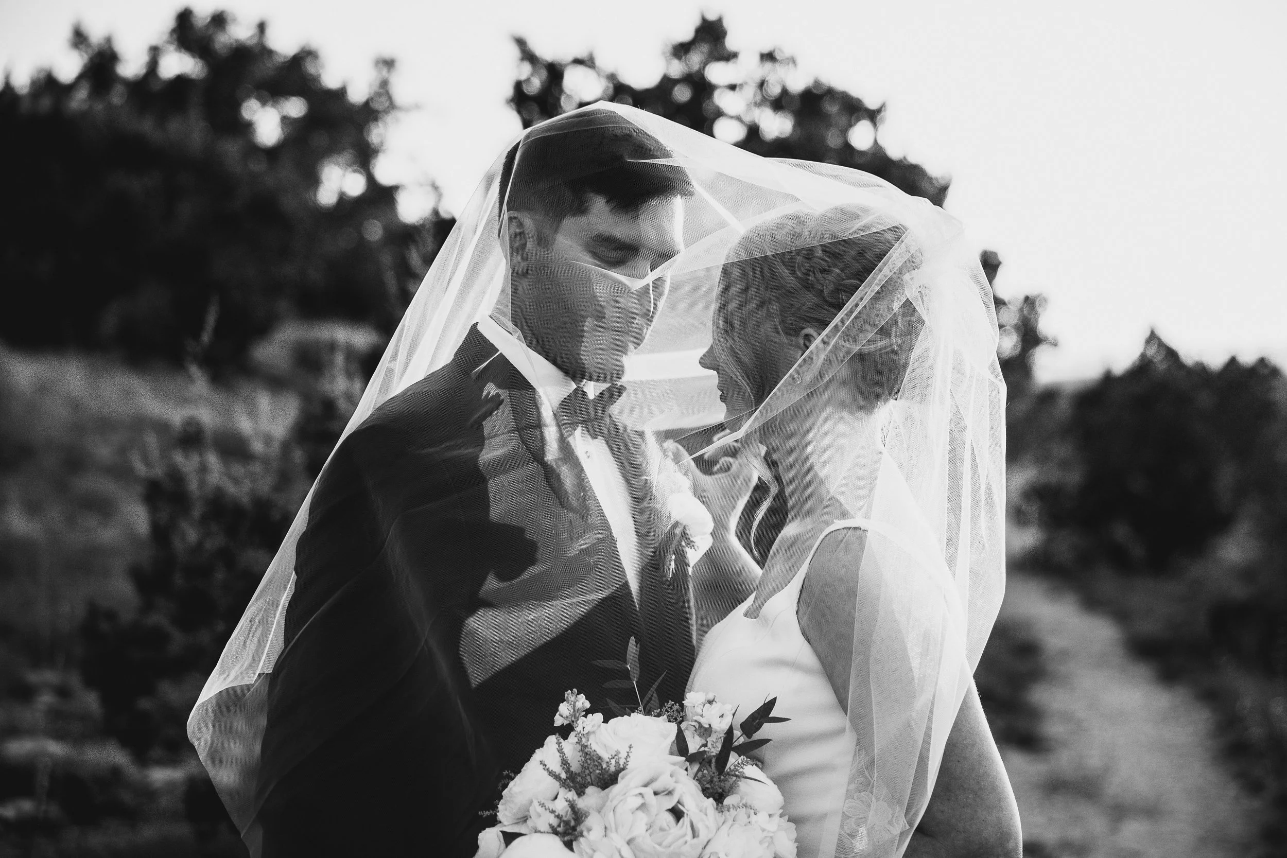 Black and white photo of a bride and groom under a veil, standing outdoors, close together, with trees in the background. The groom is holding the bride and she is holding a bouquet of flowers.