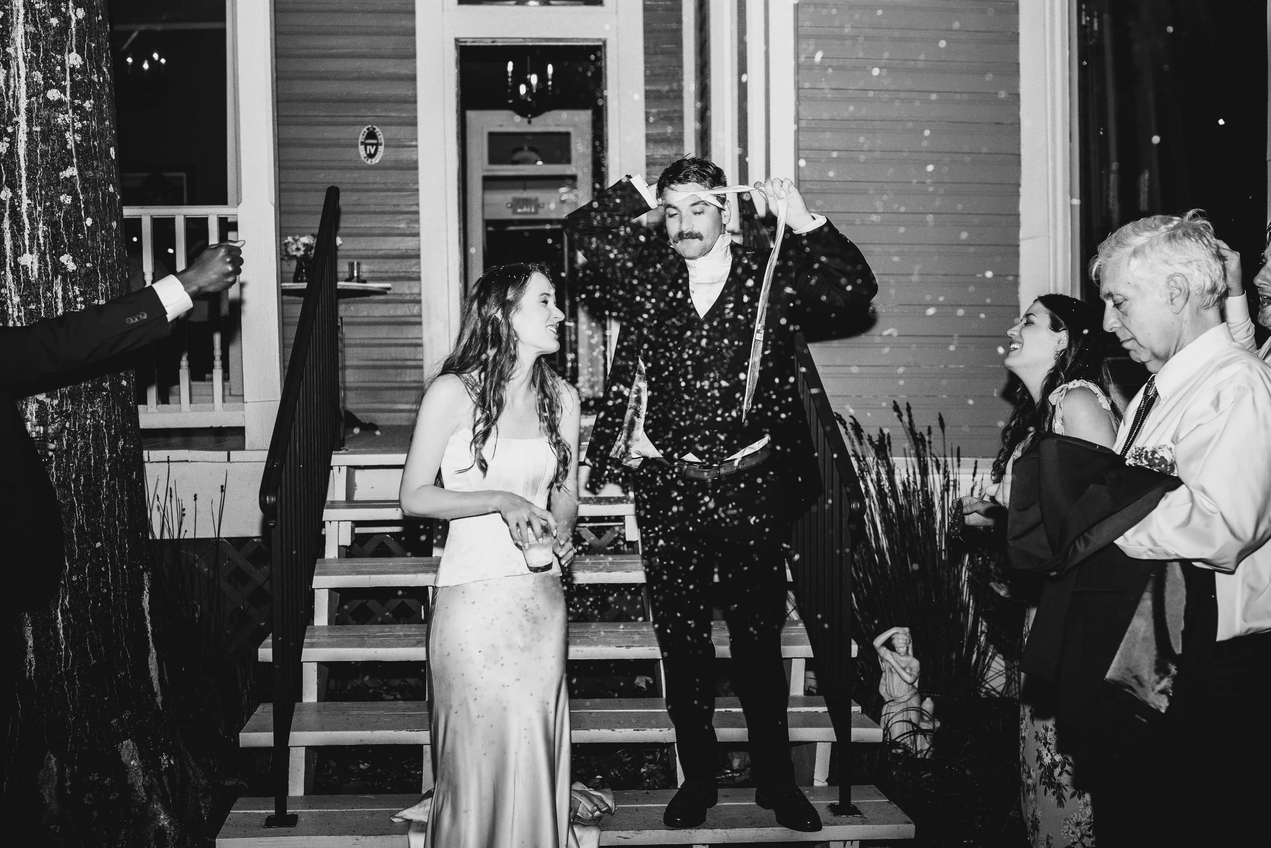 A man in a suit and a woman in a dress celebrating with guests outside a house at night, with confetti in the air.