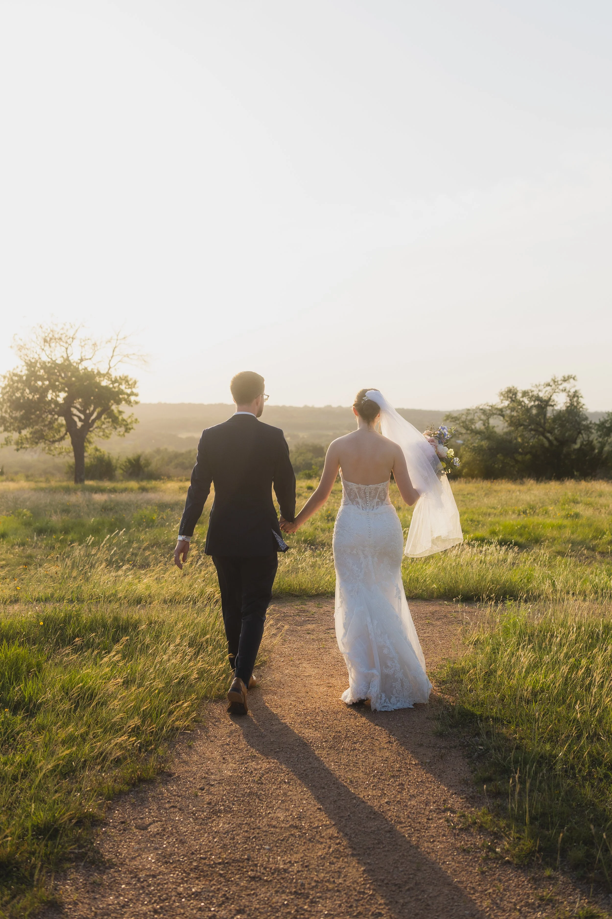 A bride and groom walk hand in hand along a dirt path in a grassy field during sunset, with trees in the distance.