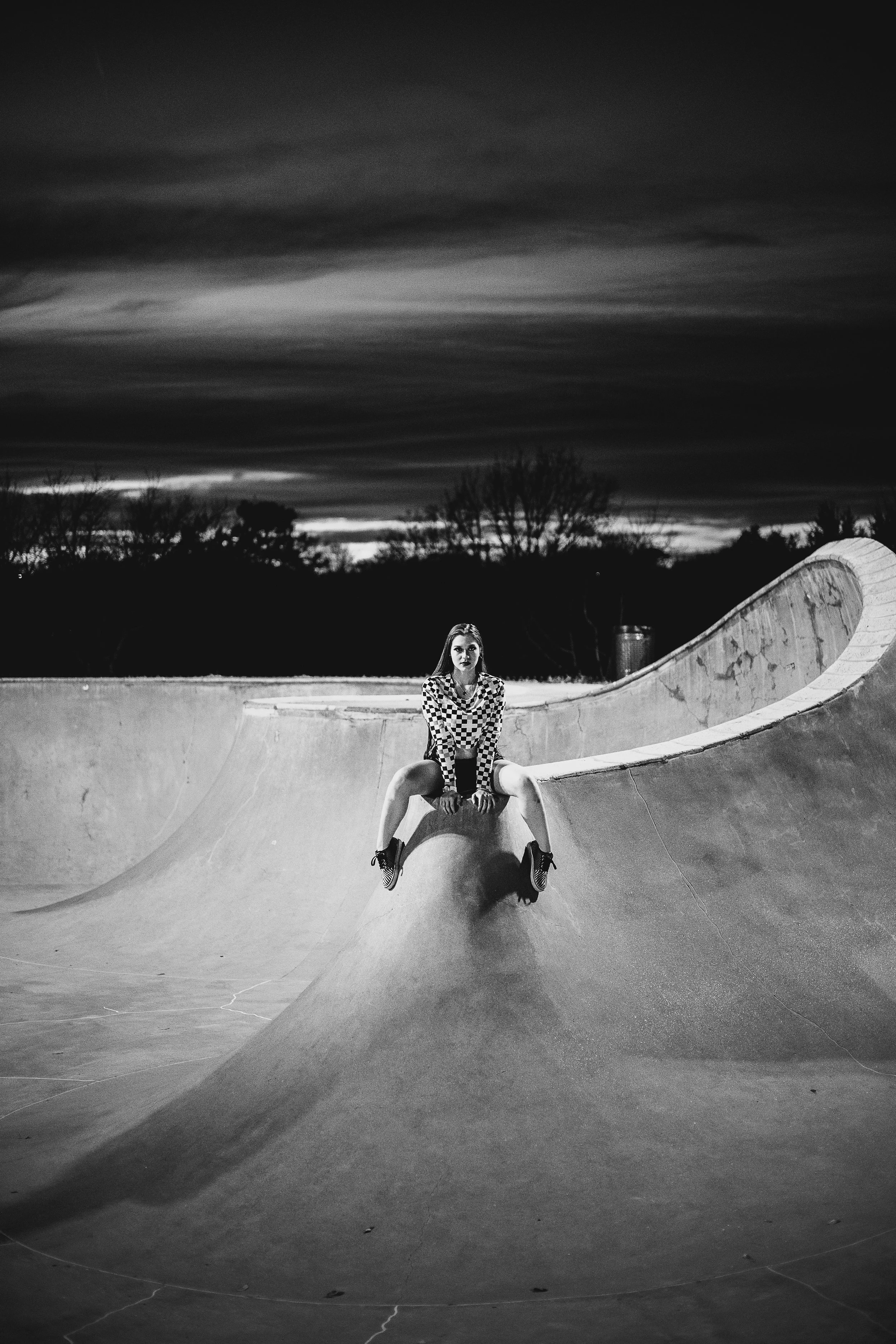 A young woman sitting on the edge of a skatepark bowl during dusk, wearing a checkered shirt and shoes, with leafless trees and a dark, cloudy sky in the background.