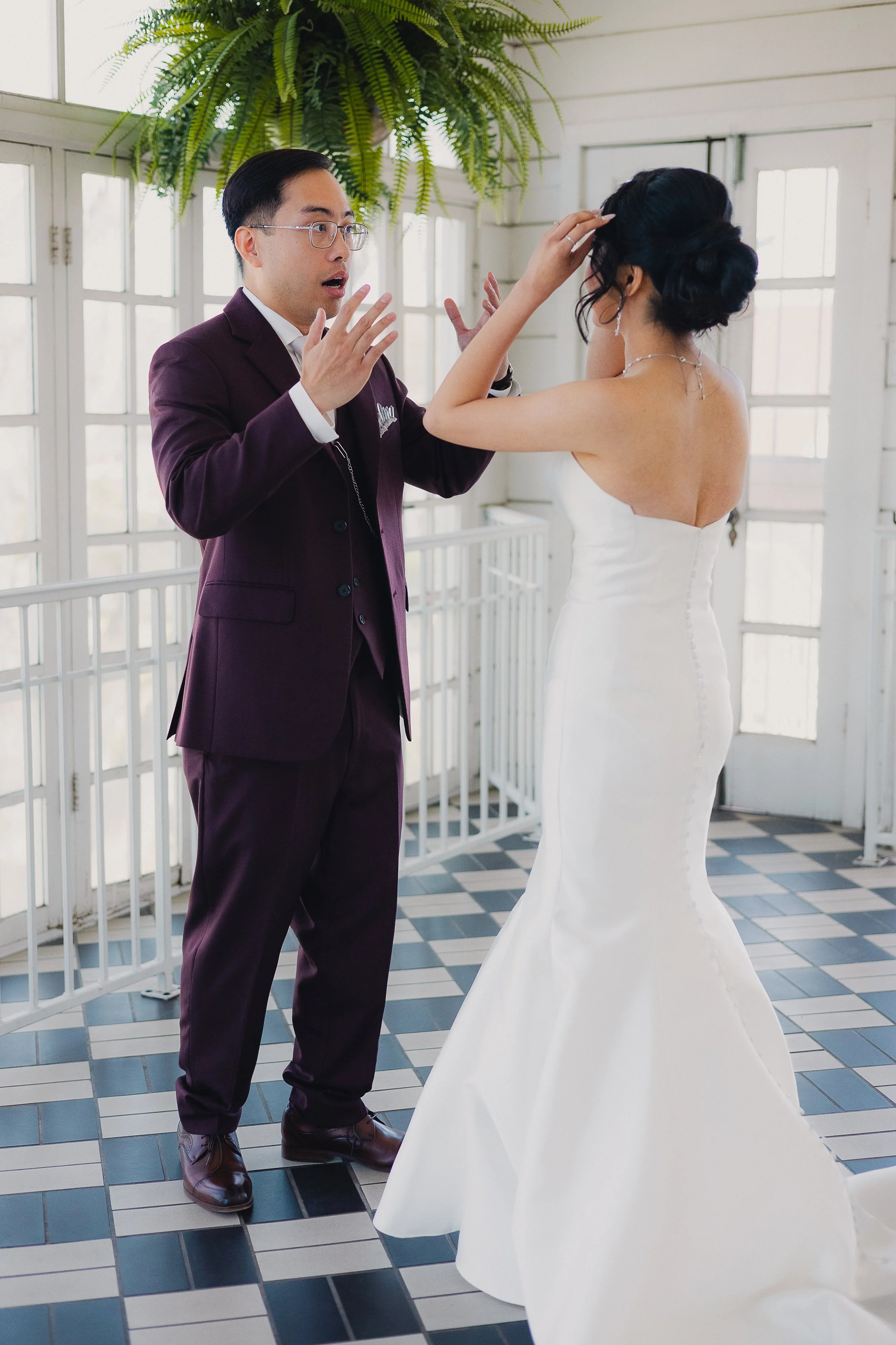 A groom in a burgundy suit with glasses is surprised as a bride in a white strapless wedding dress adjusts her hair in a bright, airy room with large windows and checkered tile flooring.