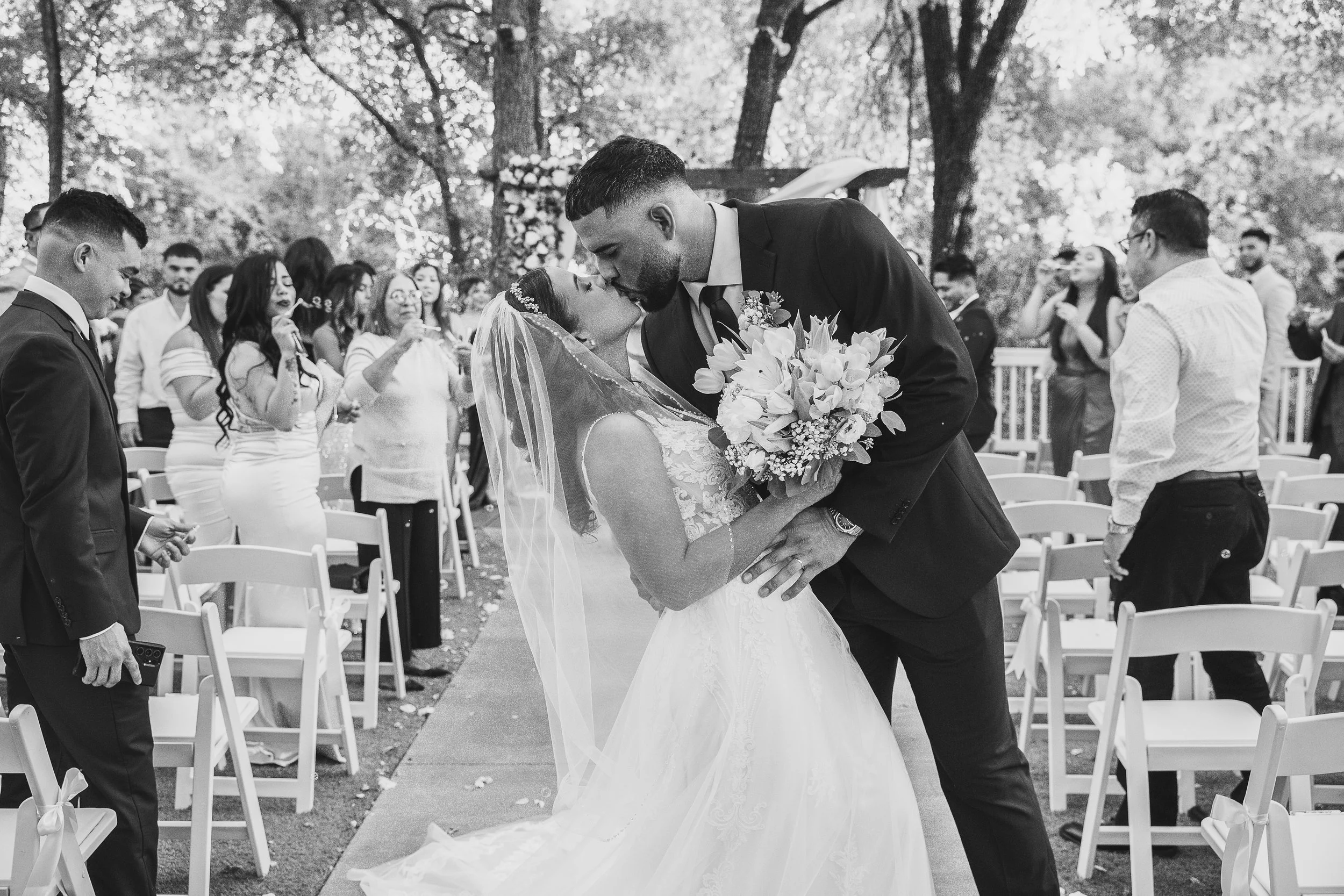 A bride and groom kissing during their outdoor wedding ceremony, with guests standing and watching.