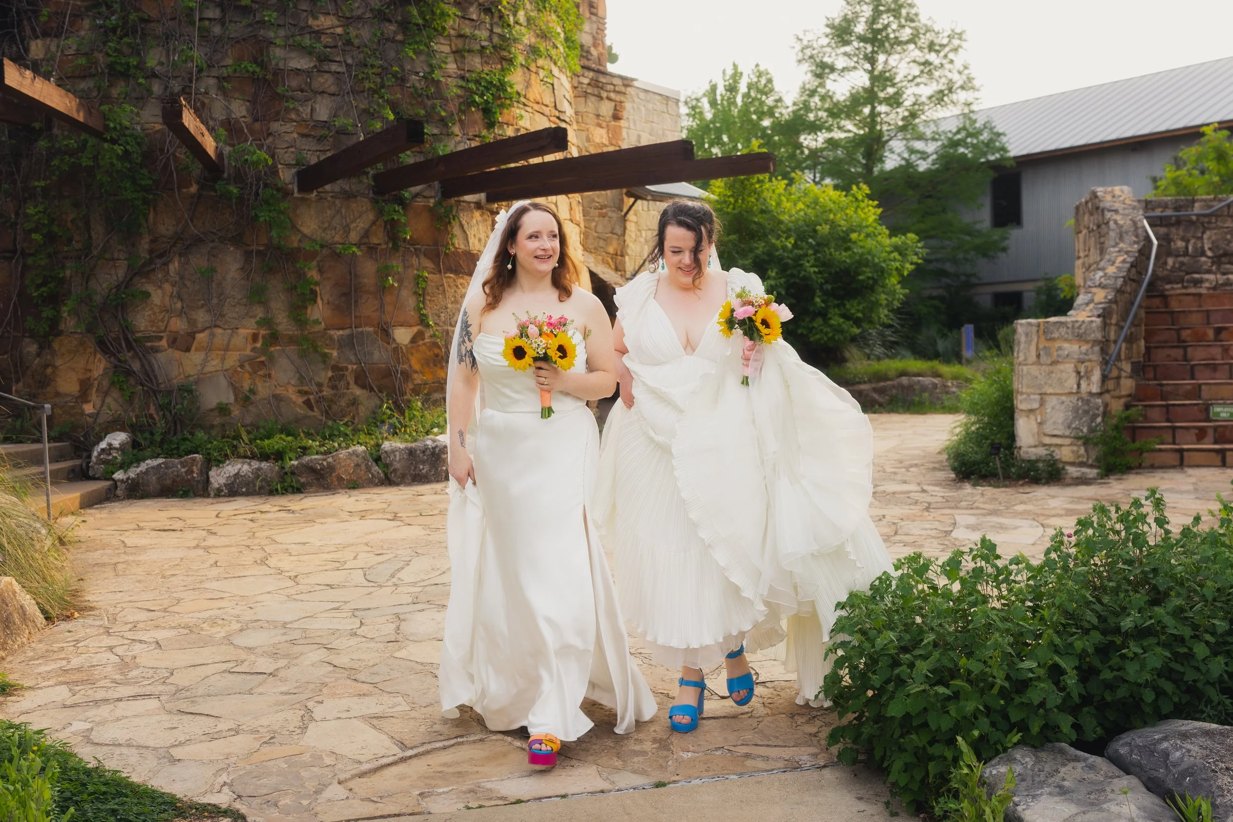 Two brides in wedding dresses walking outdoors, each holding a bouquet of sunflowers and pink flowers, with greenery and stone structures in the background.
