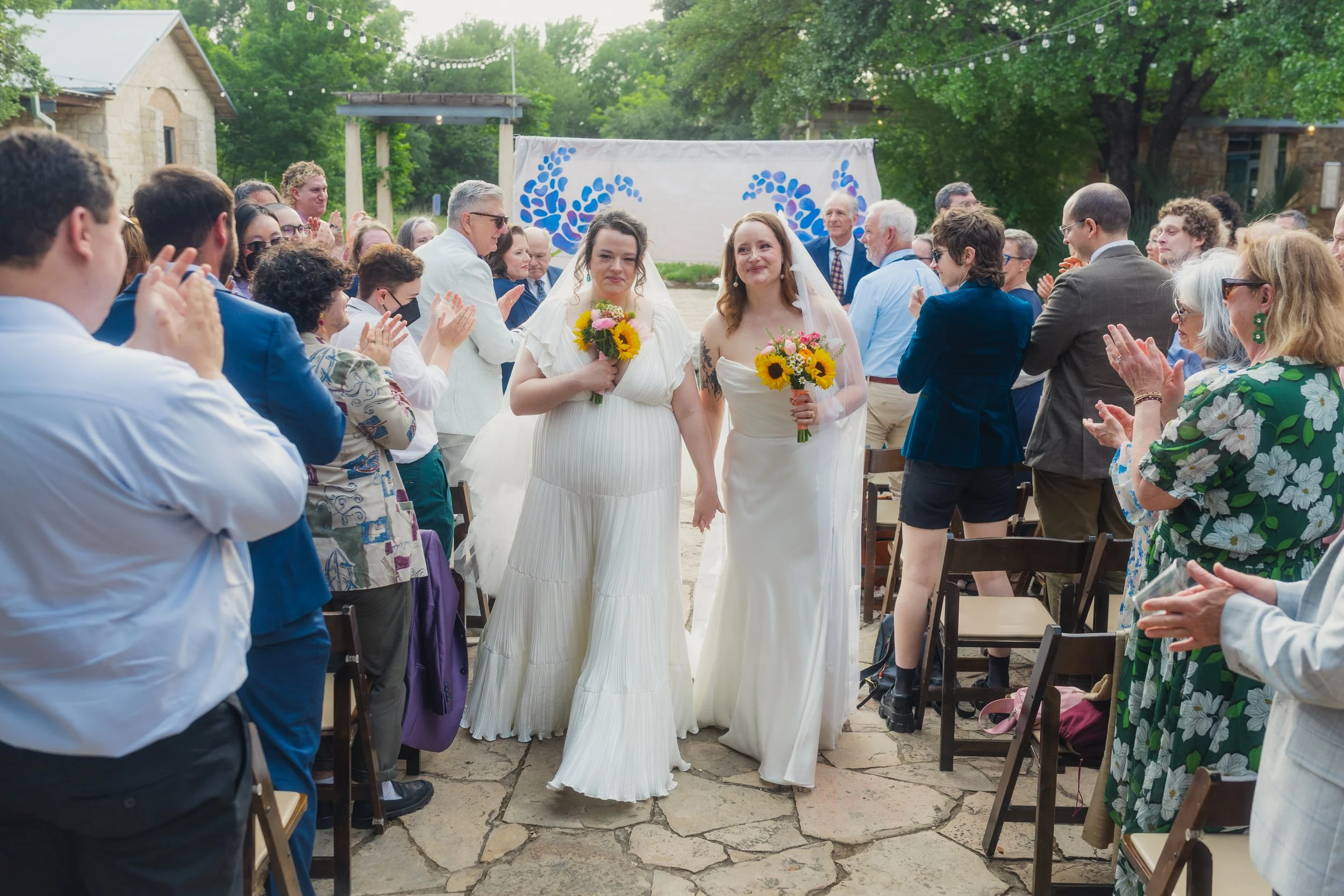 Two brides in white wedding dresses walking hand-in-hand down the aisle at an outdoor wedding ceremony, surrounded by friends and family clapping and celebrating, with greenery and string lights overhead.